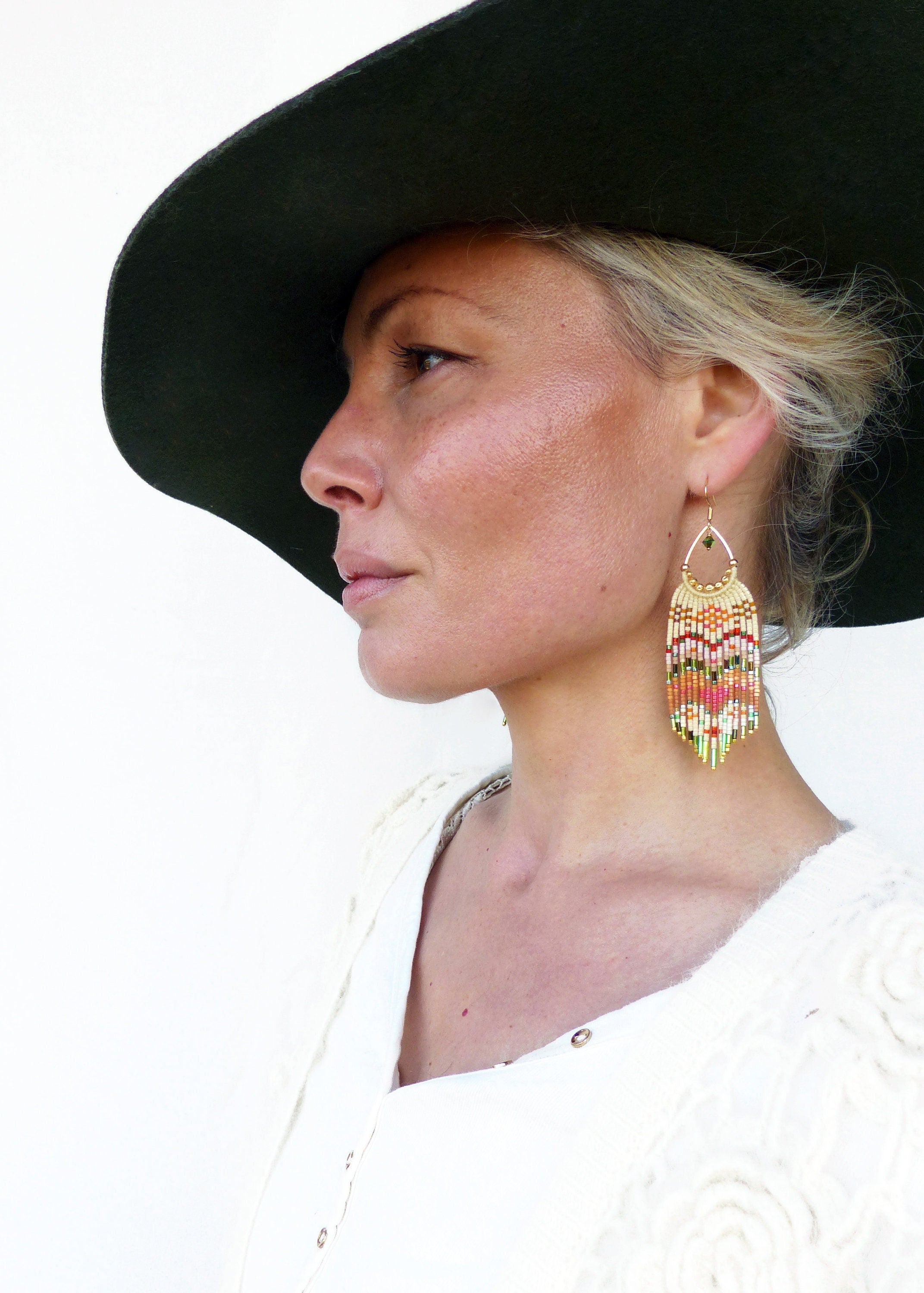 Peut inclure: Une femme portant un chapeau noir à larges bords et un pull en crochet blanc. Elle porte de longues boucles d'oreilles en perles colorées.