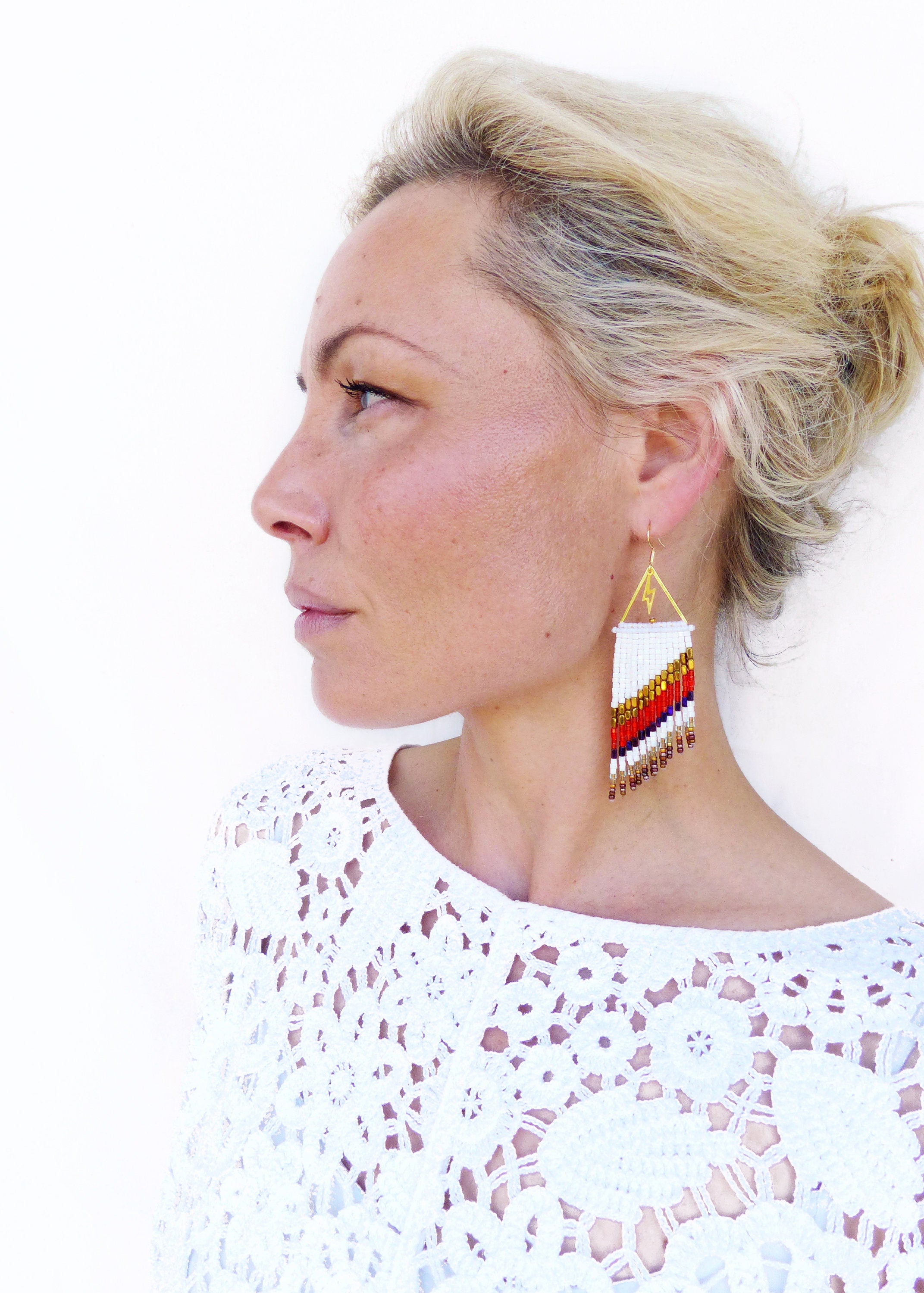 Peut inclure: Une femme portant un haut en crochet blanc avec de longs cheveux blonds. Elle porte une paire de longues boucles d'oreilles en perles avec un triangle doré en haut. Les boucles d'oreilles sont blanches avec des accents rouges, orange et bruns.