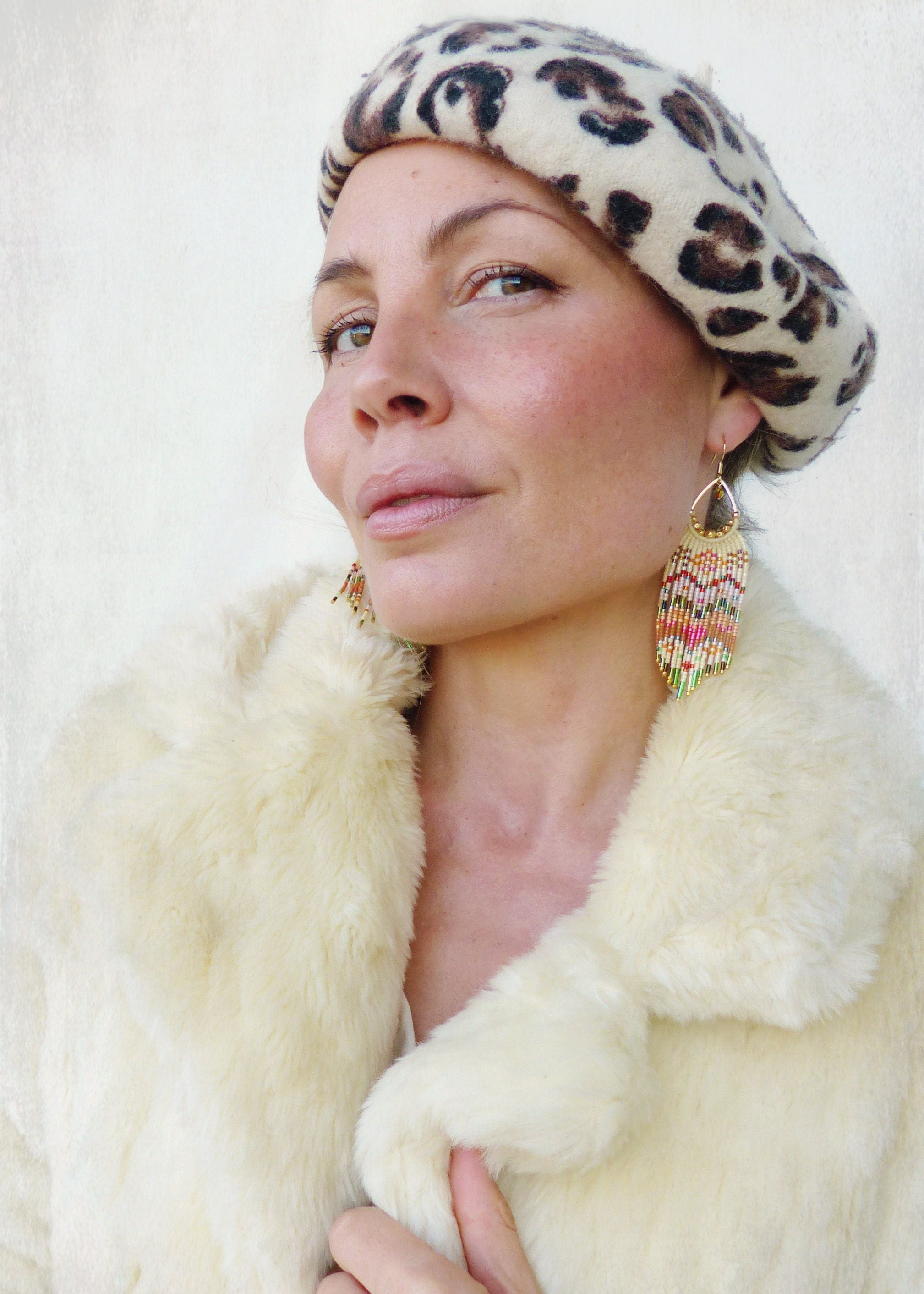 Peut inclure: Une femme portant un manteau en fausse fourrure blanc et un béret à imprimé léopard. Elle porte de longues boucles d'oreilles en perles colorées.