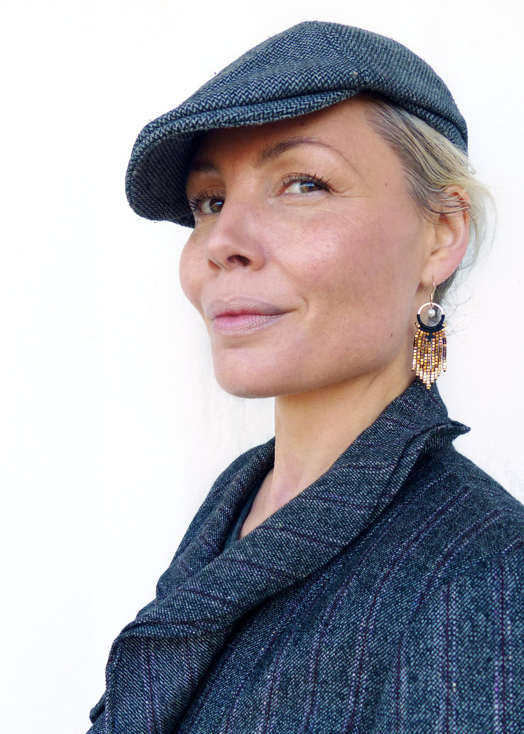 Peut inclure: Une femme portant une casquette de newsboy en tweed gris et une veste en tweed gris assortie. Elle porte des boucles d'oreilles en or avec des perles brunes et noires.