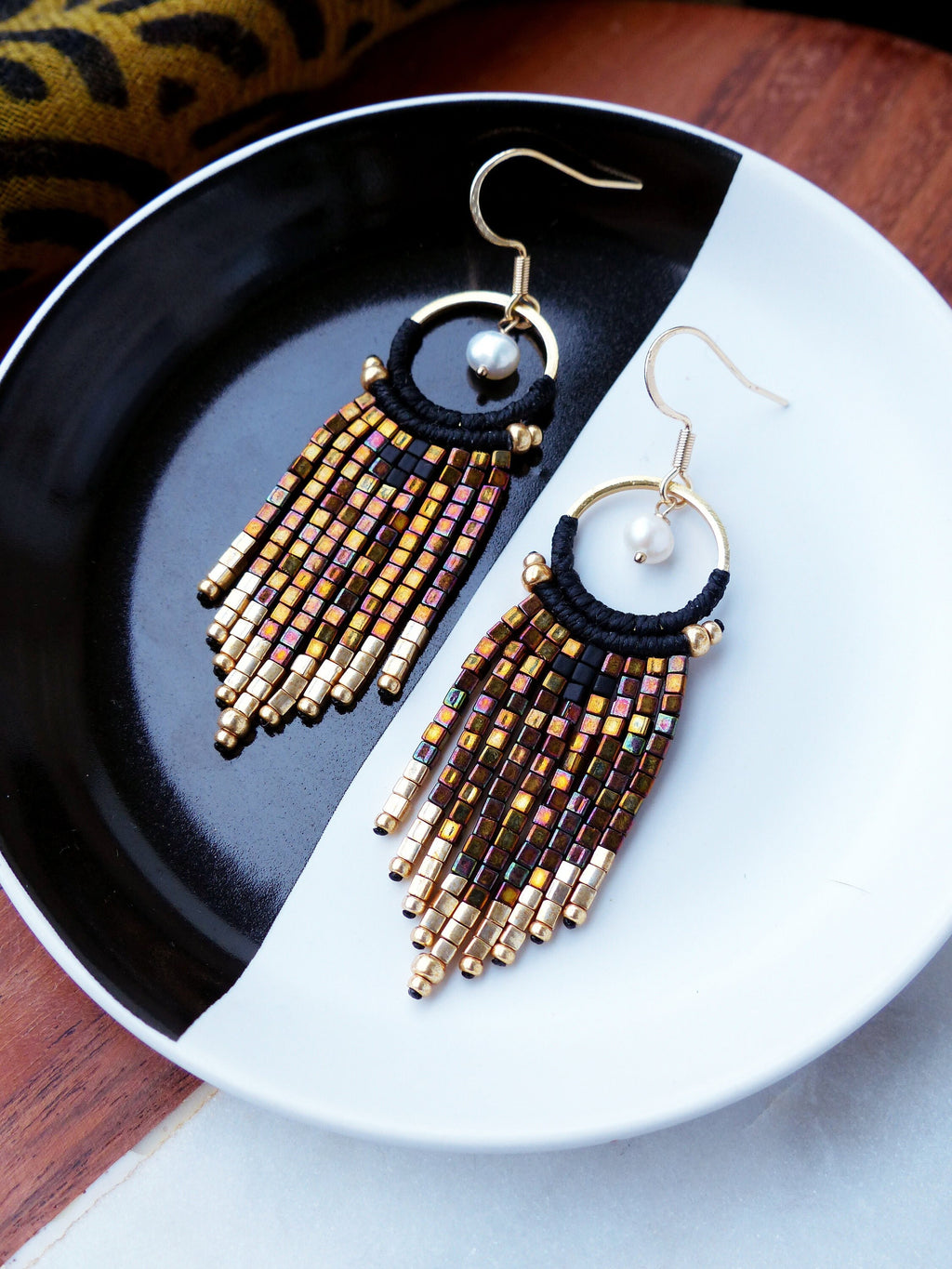 Peut inclure: Une paire de boucles d'oreilles créoles dorées avec du fil noir et des perles dorées irisées qui pendent du bas. Chaque boucle d'oreille a un petit charme de perle blanche.