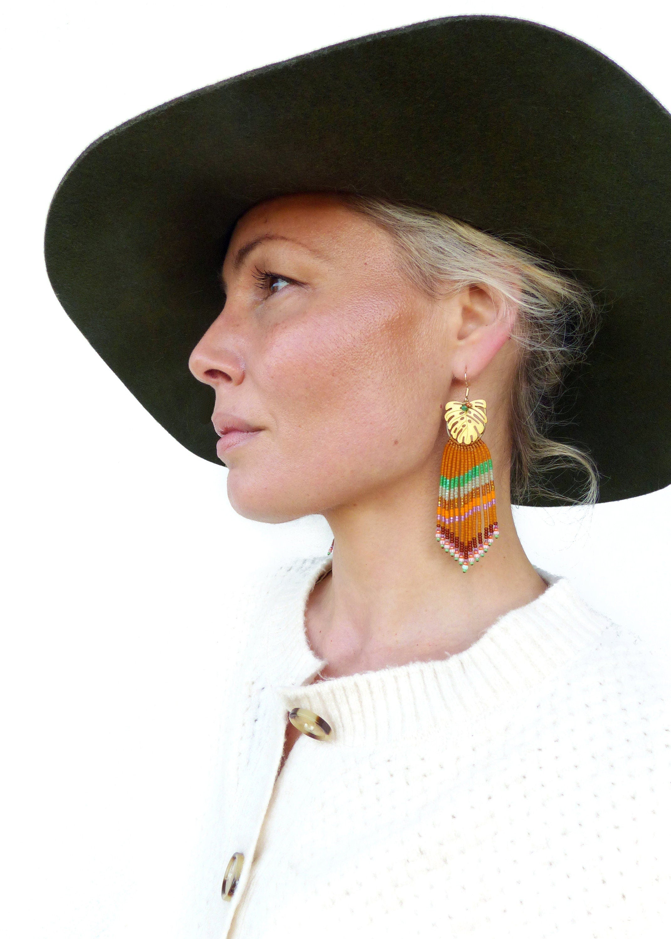 Peut inclure: Boucles d'oreilles dorées en forme de feuille avec des franges de perles colorées. La personne porte un chapeau vert foncé et un cardigan blanc. Les franges de perles sont orange, vertes et roses.