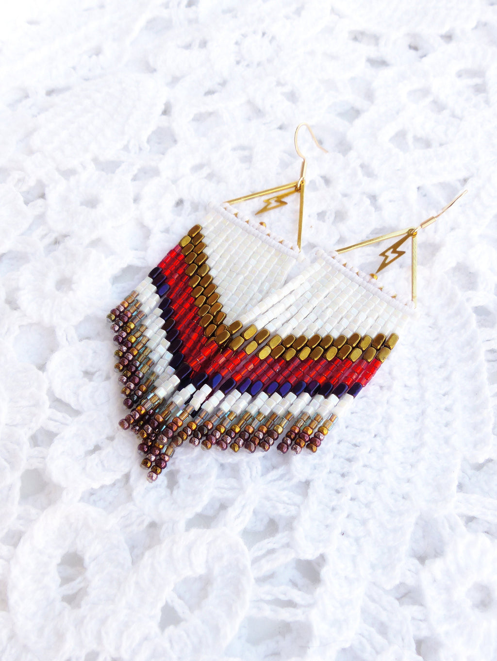 Peut inclure: Une paire de boucles d'oreilles en perles blanches, rouges, dorées et violettes avec un charme éclair doré. Les boucles d'oreilles sont suspendues à des crochets dorés.
