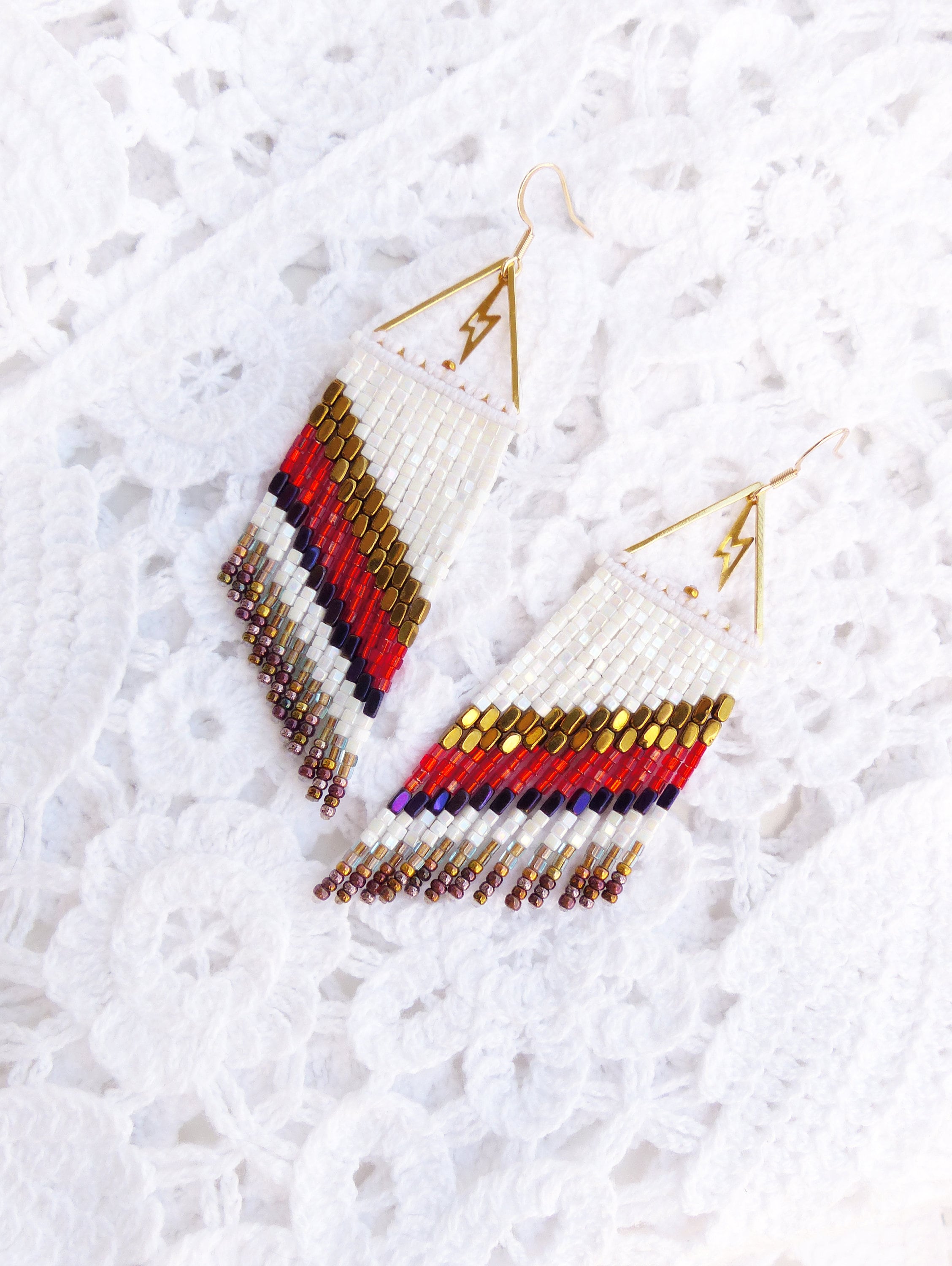 Peut inclure: Une paire de longues boucles d'oreilles en perles avec un motif blanc, rouge, or et violet. Les boucles d'oreilles sont en forme de triangle avec un motif d'éclair en haut.