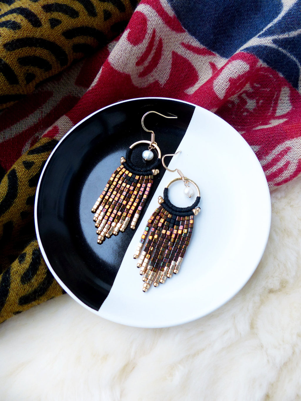 Peut inclure: Une paire de boucles d'oreilles en perles dorées et brunes avec un fil noir et un accent de perle. Les boucles d'oreilles sont présentées sur une assiette noire et blanche.