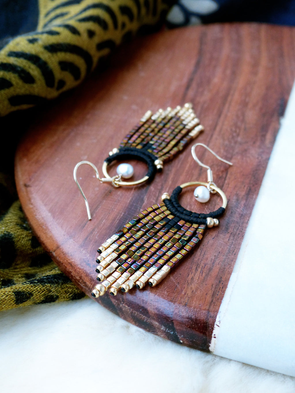 Peut inclure: Une paire de boucles d'oreilles en perles dorées et noires avec un accent de perle. Les boucles d'oreilles sont suspendues à une surface en bois.