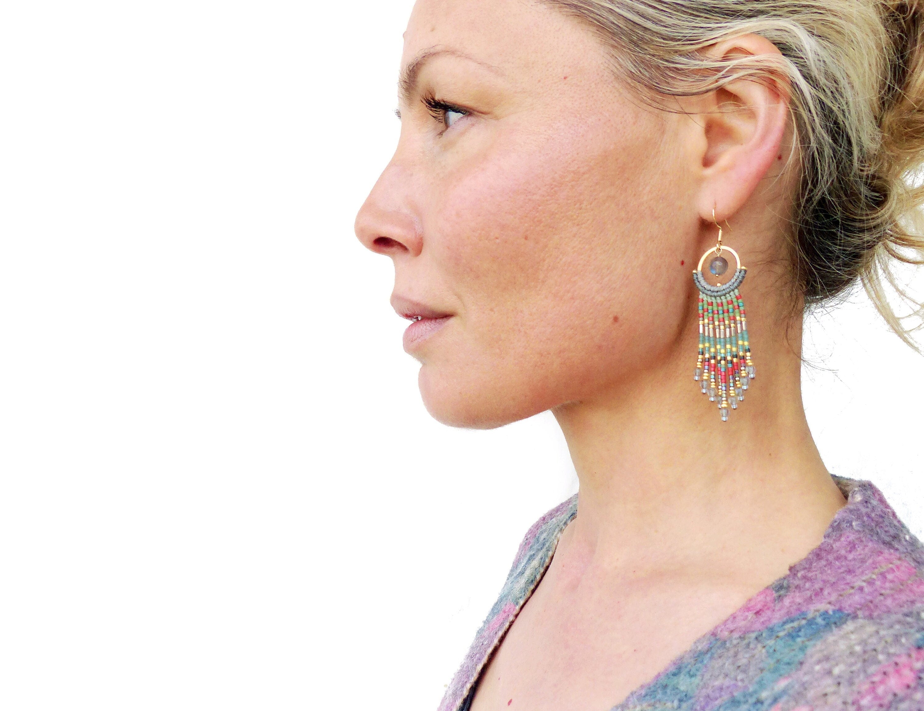 Peut inclure: Une femme portant une chemise à motifs violets et gris avec de longues boucles d'oreilles en perles. Les boucles d'oreilles sont dorées et ont un design circulaire avec une pierre grise au centre. Les boucles d'oreilles sont ornées de perles colorées, notamment rouges, vertes et bleues.