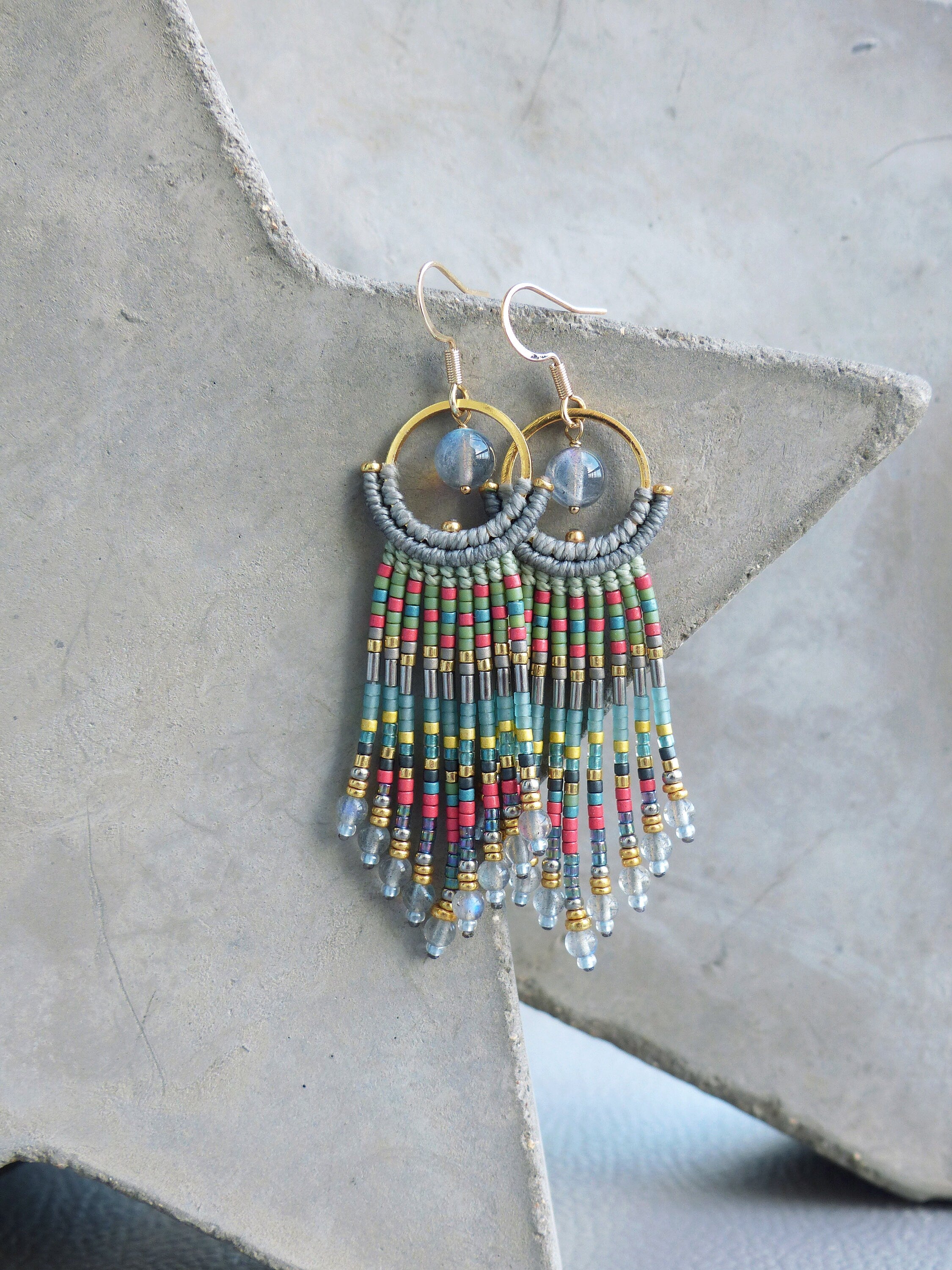 Peut inclure: Une paire de longues boucles d'oreilles en perles avec un anneau en or et un pendentif en pierre bleue. Les boucles d'oreilles présentent une frange multicolores de perles dans les tons de vert, bleu, rouge et or.