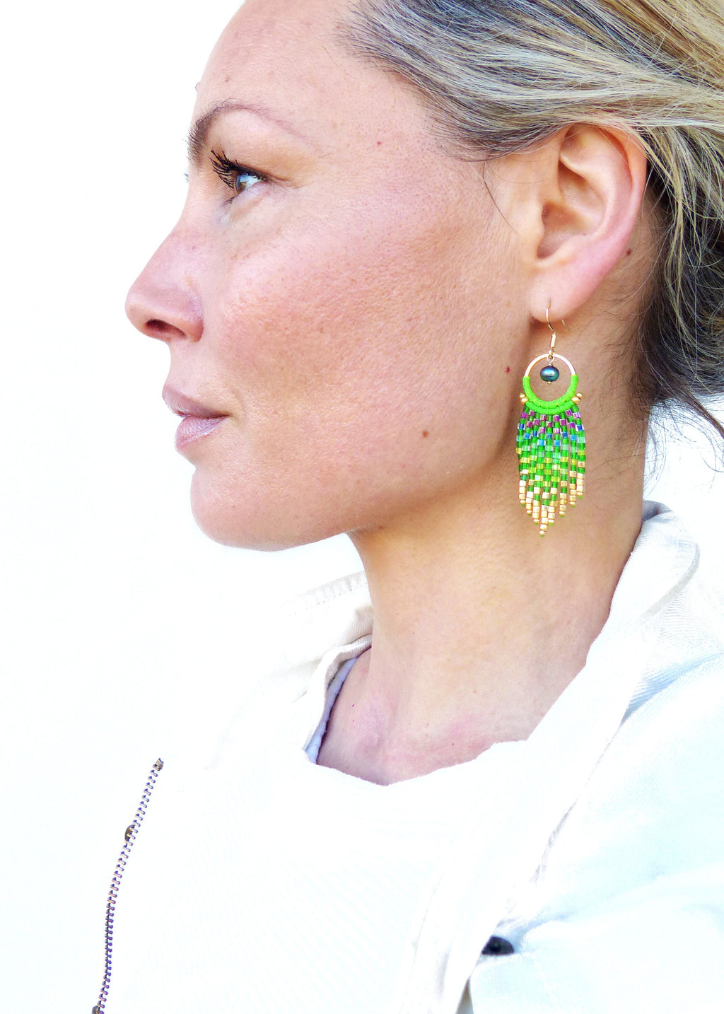 Peut inclure: Une femme porte des boucles d'oreilles en perles. Les boucles d'oreilles ont un cercle doré avec un motif de perles vertes et violettes et une frange de perles vertes, jaunes et dorées. Un accessoire coloré.