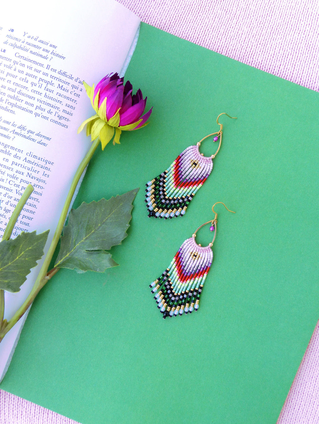Peut inclure: Une paire de boucles d'oreilles en perles colorées avec un design en frange. Les boucles d'oreilles sont faites de perles noires, blanches, vertes, rouges et roses. Les boucles d'oreilles sont suspendues à des crochets dorés.