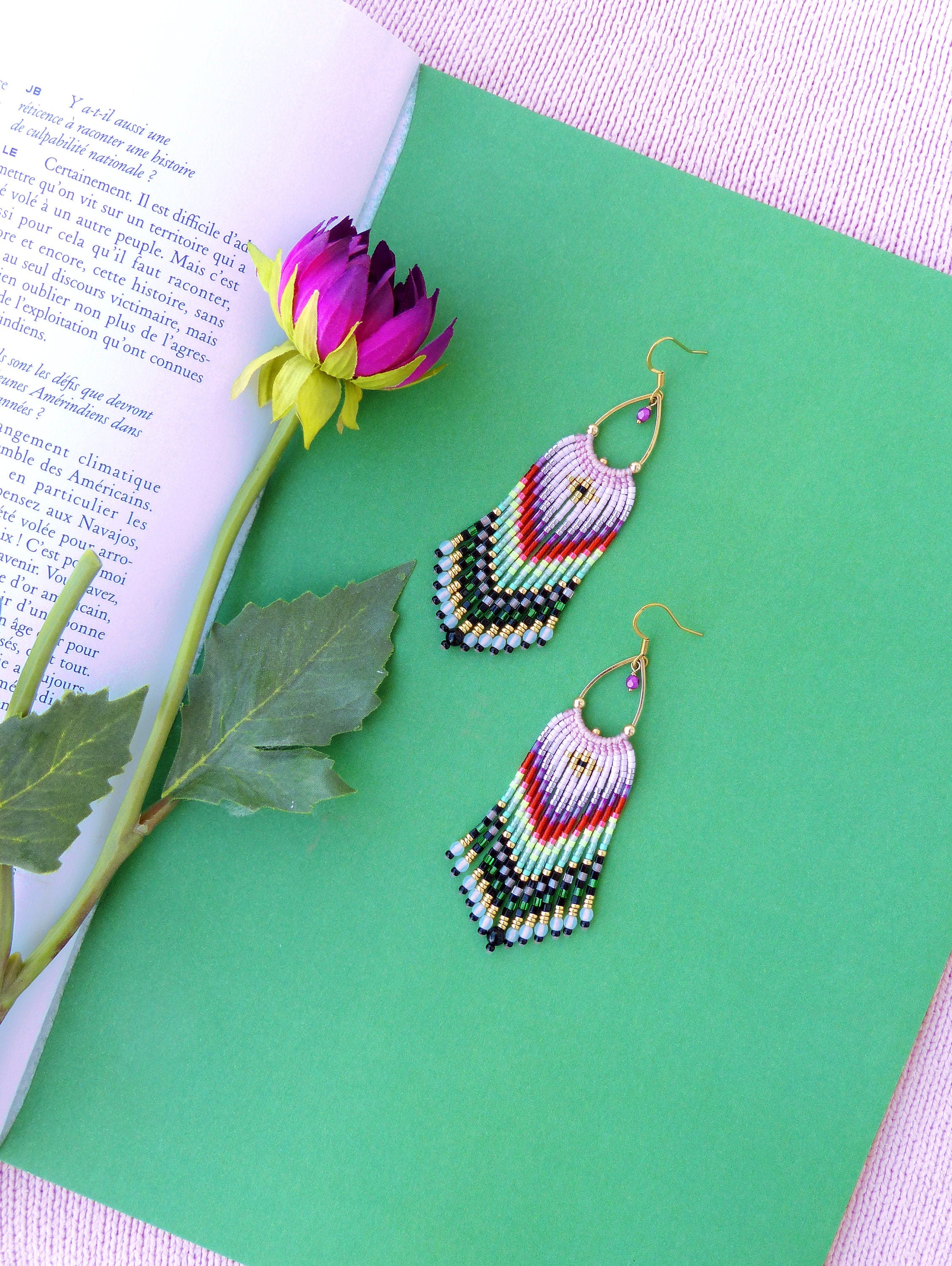 Peut inclure: Une paire de boucles d'oreilles en perles colorées avec un design en frange. Les boucles d'oreilles sont faites de perles noires, blanches, vertes, rouges et roses. Les boucles d'oreilles sont suspendues à des crochets dorés.