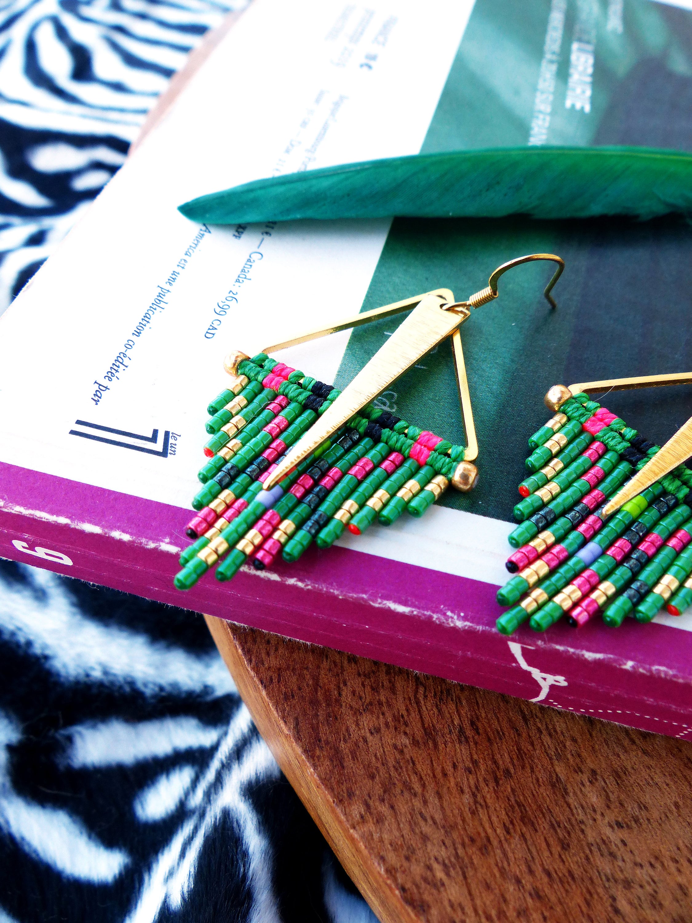 Peut inclure: Une paire de boucles d'oreilles triangulaires dorées avec des perles vertes, roses, noires et dorées. Les boucles d'oreilles sont suspendues à un livre.