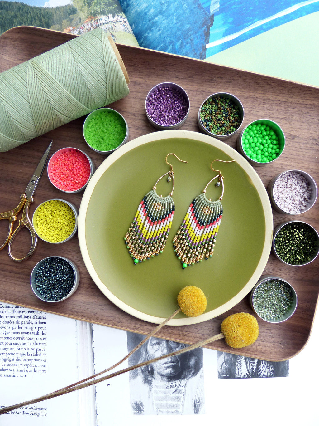 Peut inclure: Une paire de boucles d'oreilles en perles dorées avec un design en frange. Les boucles d'oreilles sont faites de perles vertes, jaunes, blanches et noires. Les boucles d'oreilles sont présentées sur une assiette verte avec un plateau en bois en arrière-plan.