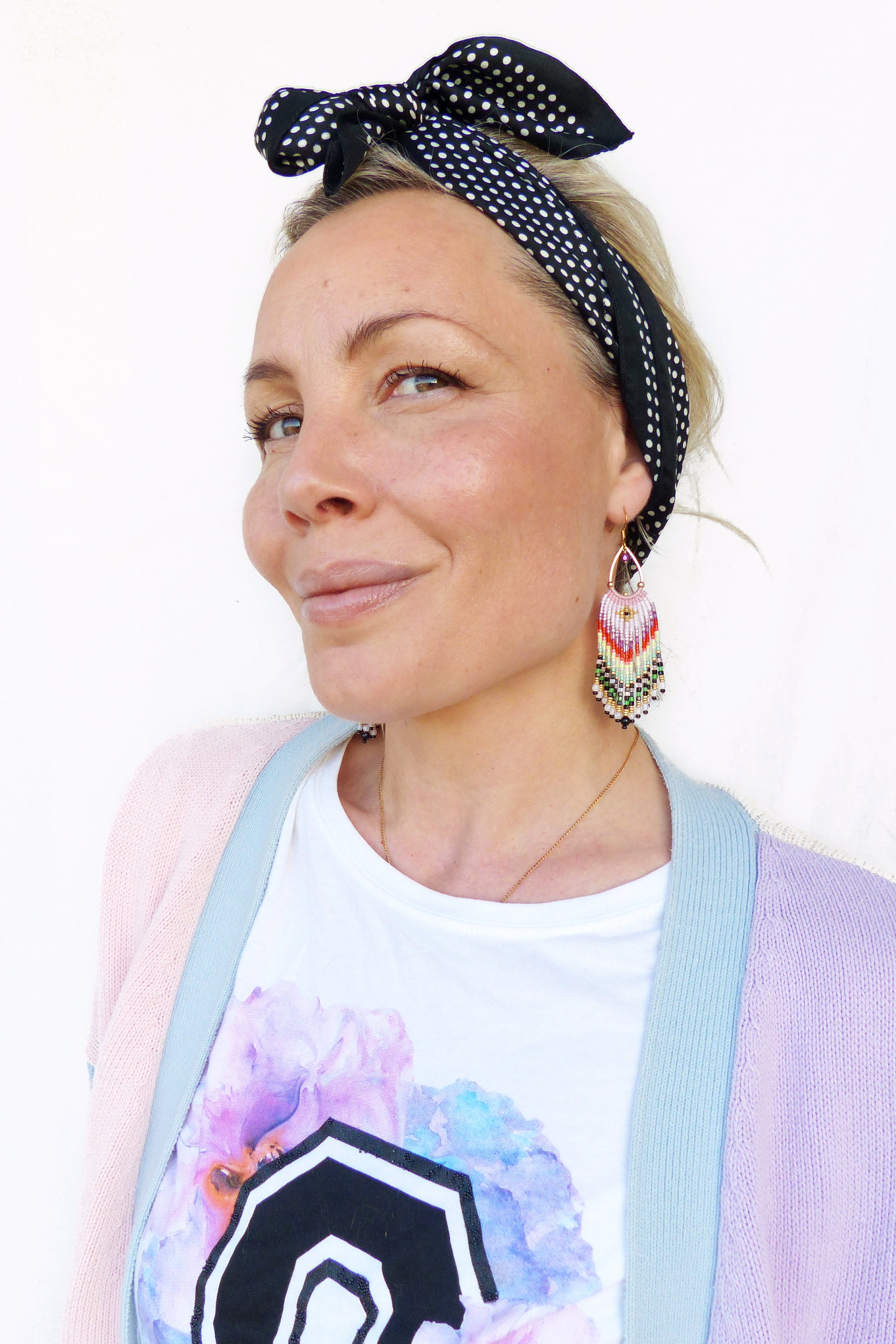 Peut inclure: Une femme portant un t-shirt blanc avec un motif floral aquarelle, un cardigan rose clair avec une bordure bleue et un bandeau à pois noir et blanc. Elle porte de longues boucles d'oreilles en perles de couleur rose, verte et orange.