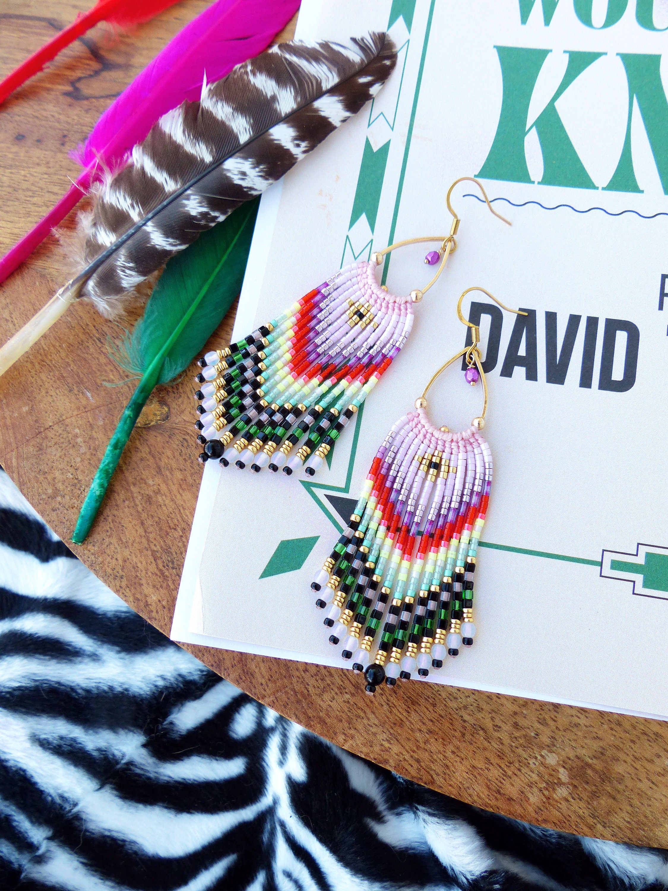 Peut inclure: Une paire de boucles d'oreilles en perles colorées avec un design en frange. Les boucles d'oreilles sont faites de perles roses, violettes, rouges, vertes, noires et blanches. Les boucles d'oreilles sont suspendues à des crochets dorés.