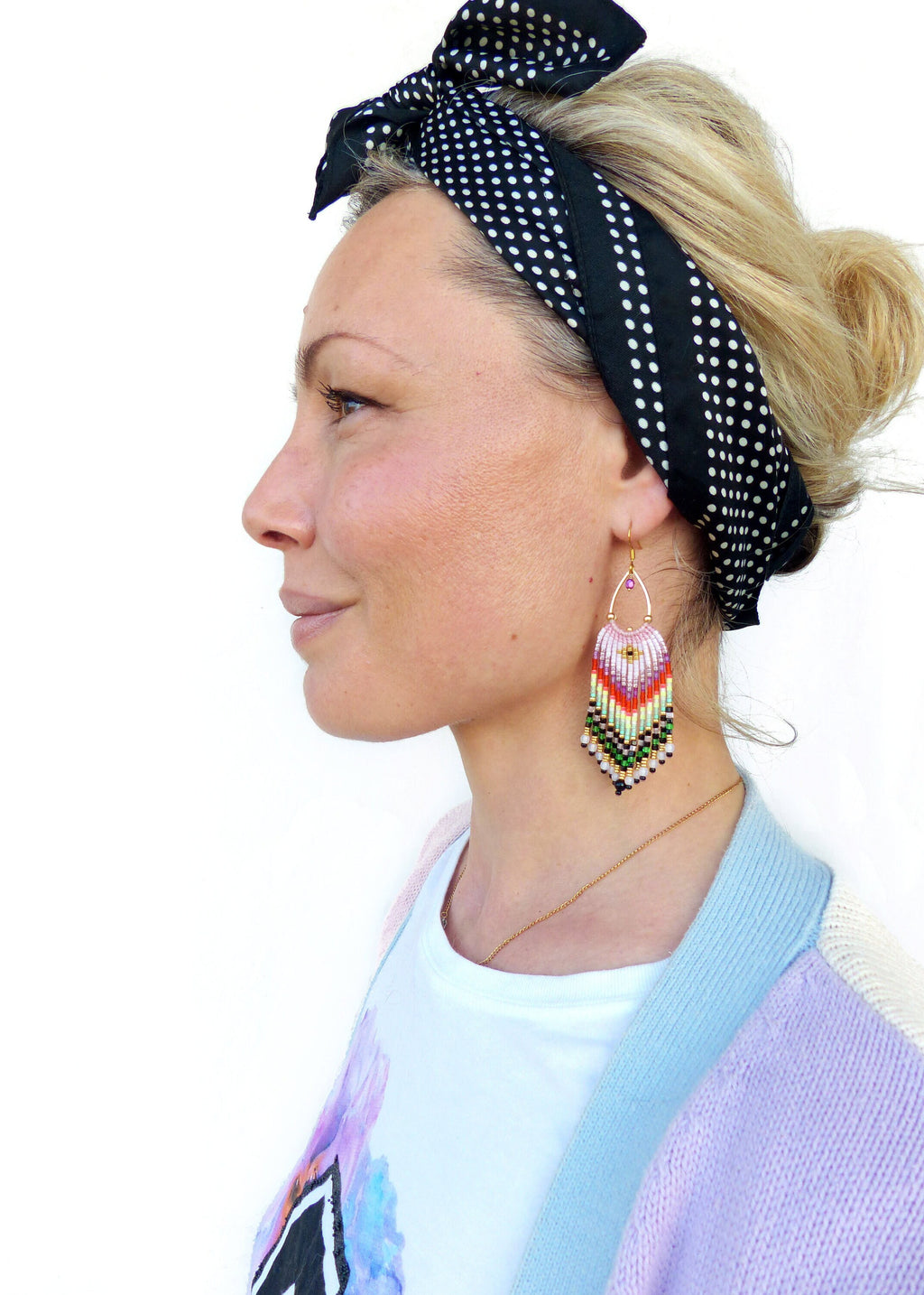 Peut inclure: Une femme portant un t-shirt blanc avec un motif coloré, un cardigan bleu clair et violet et un bandeau à pois noirs et blancs. Elle porte de longues boucles d'oreilles en perles colorées.