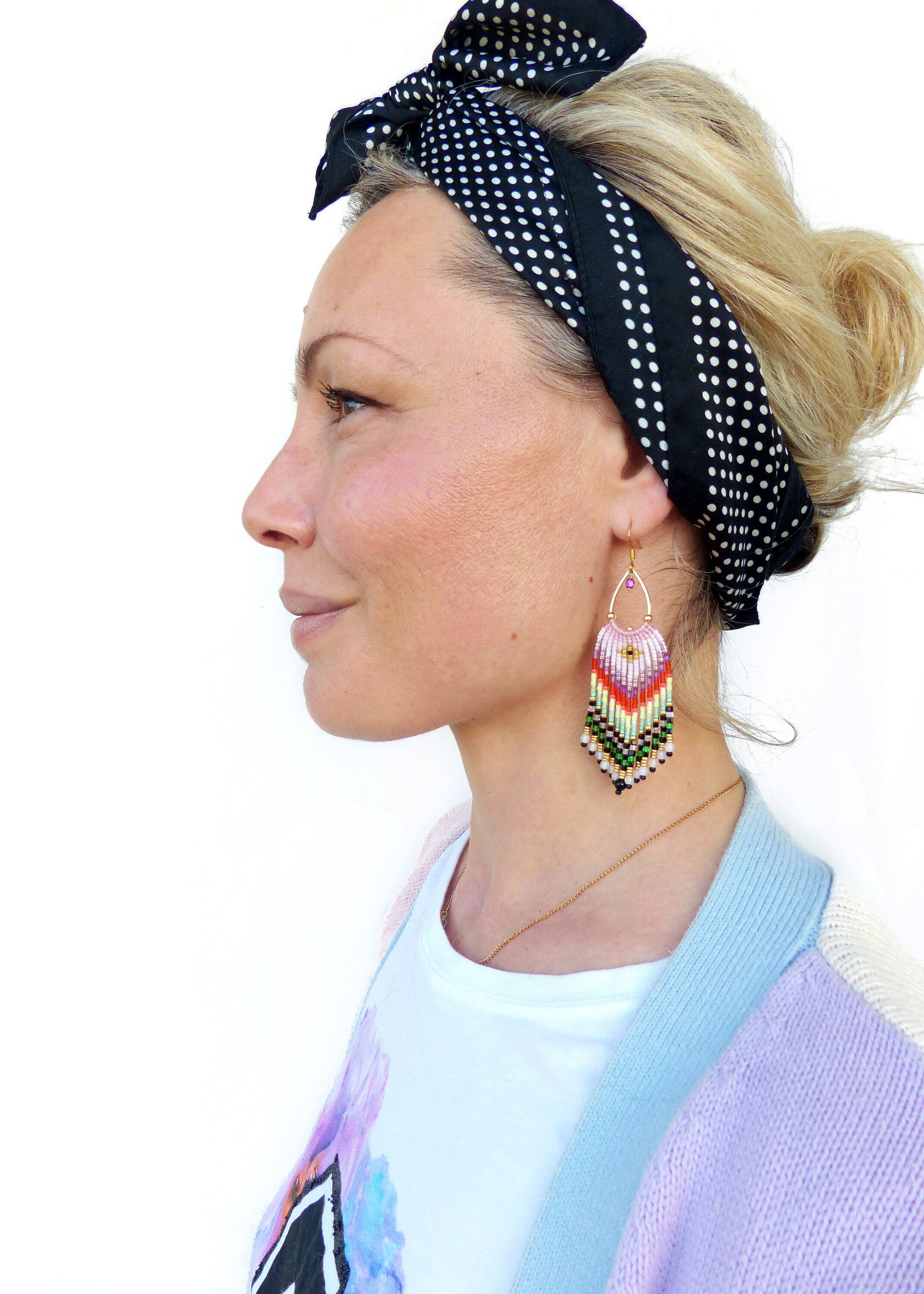 Peut inclure: Une femme portant un t-shirt blanc avec un motif coloré, un cardigan bleu clair et violet et un bandeau à pois noirs et blancs. Elle porte de longues boucles d'oreilles en perles colorées.