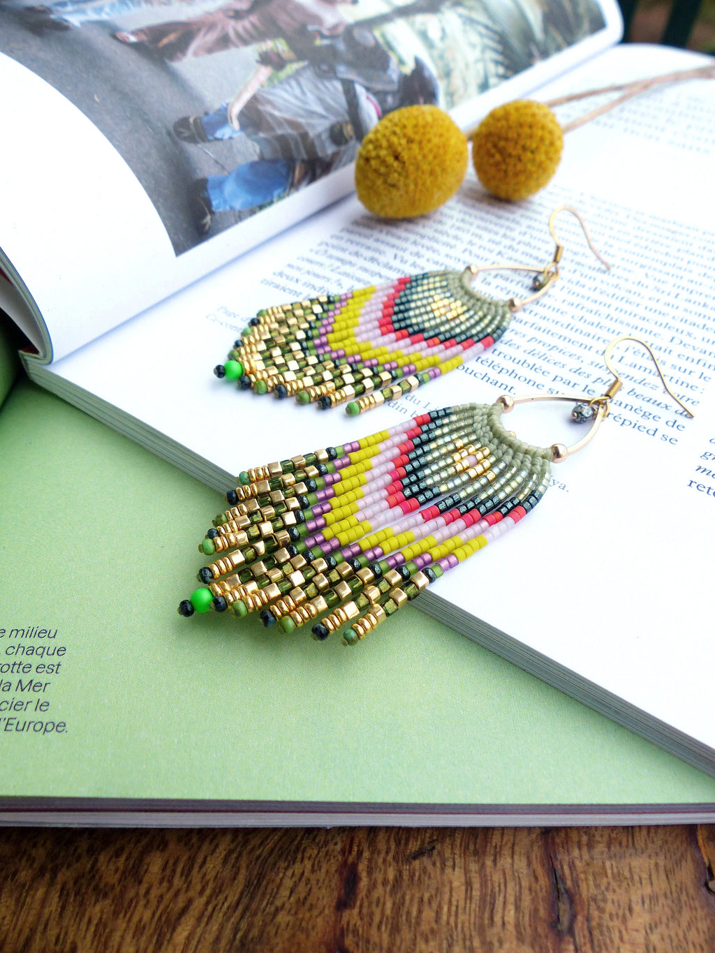 Peut inclure: Une paire de longues boucles d'oreilles en perles colorées avec un schéma de couleurs vert, jaune, rose et or. Les boucles d'oreilles sont suspendues à un livre.