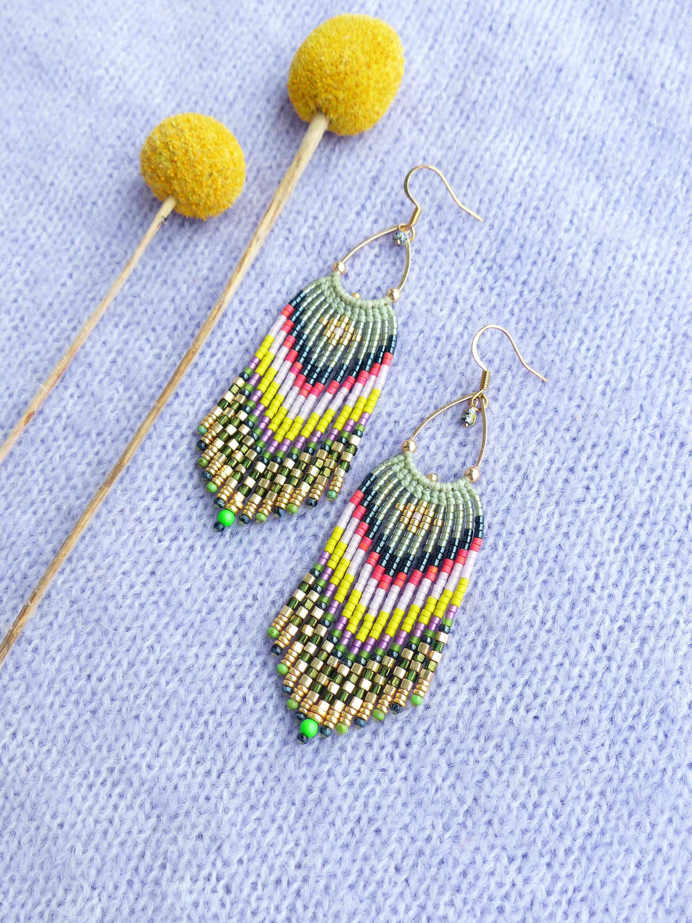 Peut inclure: Une paire de longues boucles d'oreilles en perles avec un motif géométrique dans des tons de vert, jaune, rose et noir. Les boucles d'oreilles sont suspendues à des crochets dorés.