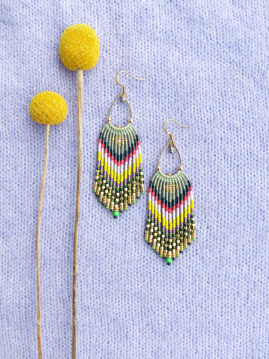 Peut inclure: Une paire de longues boucles d'oreilles en perles colorées avec un motif vert, jaune, blanc, rose et noir. Les boucles d'oreilles sont suspendues à des crochets dorés et se trouvent sur un fond en tricot de couleur violet clair.