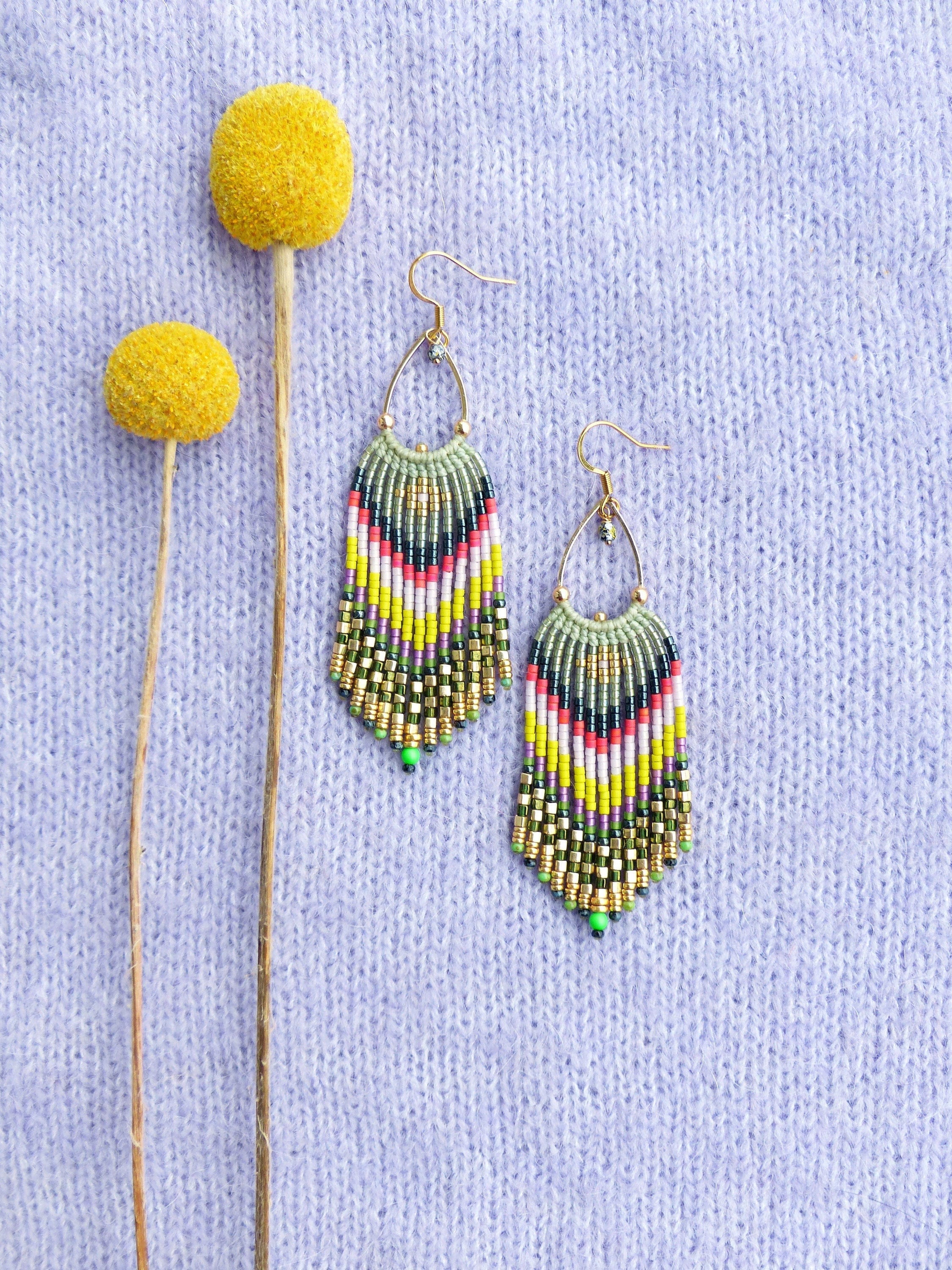 Peut inclure: Une paire de longues boucles d'oreilles en perles colorées avec un motif vert, jaune, blanc, rose et noir. Les boucles d'oreilles sont suspendues à des crochets dorés et se trouvent sur un fond en tricot de couleur violet clair.