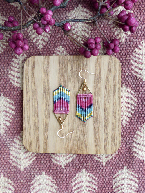 Peut inclure: Une paire de boucles d'oreilles en perles rose, bleu et or avec un motif géométrique. Les boucles d'oreilles sont suspendues à une surface en bois.