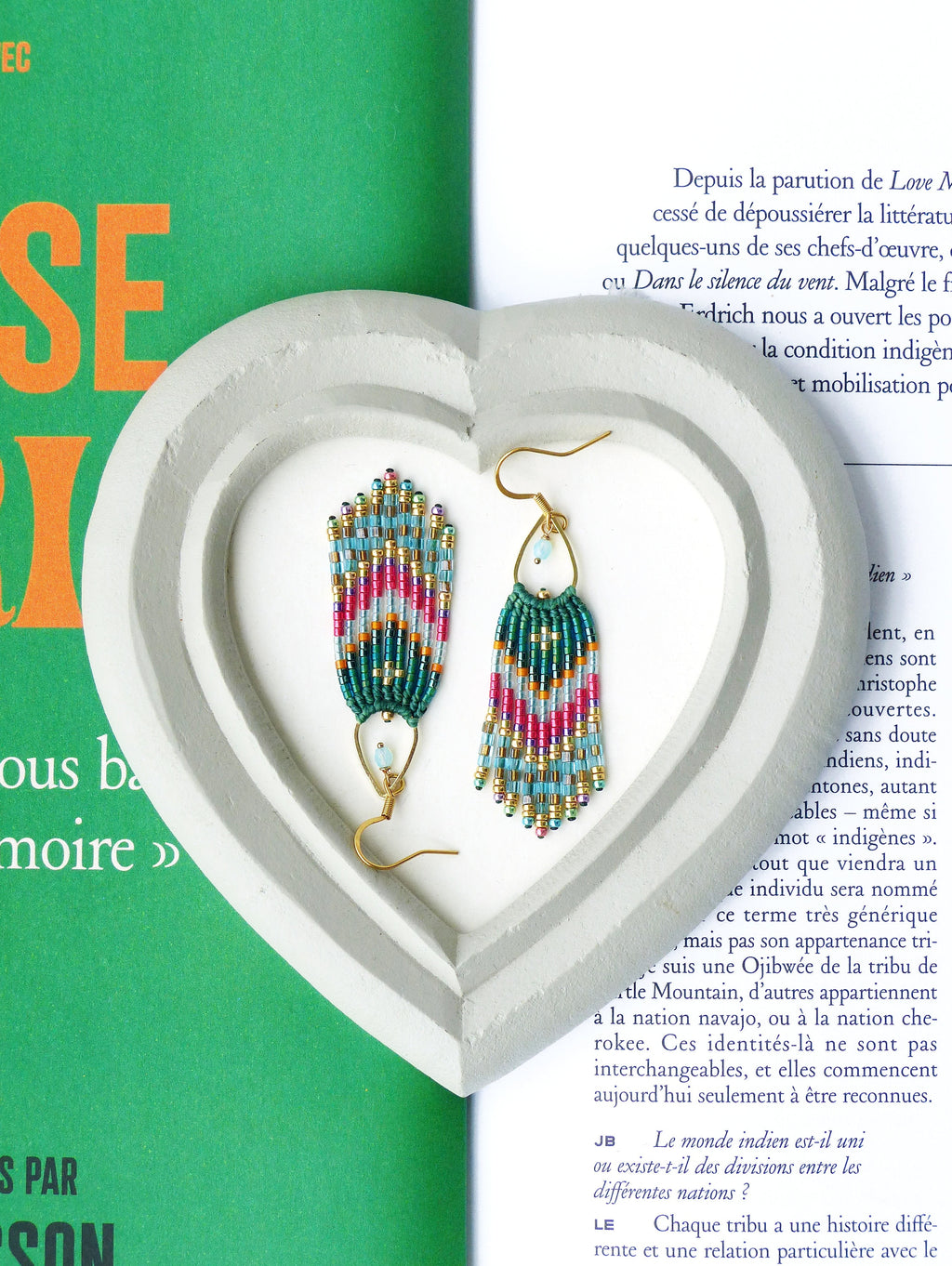 Peut inclure: Une paire de boucles d'oreilles en perles turquoise, rose et or, présentées sur un présentoir en forme de cœur. Les boucles d'oreilles sont faites de petites perles et ont un design en forme de frange. Le texte sur la page dit "Depuis la parution de Love A cessé de dépoussiérer la littérature quelques-uns de ses chefs-d'œuvre, cu Dans le silence du vent. Malgré le f drich nous a ouvert les po- la condition indigèn t mobilisation p"