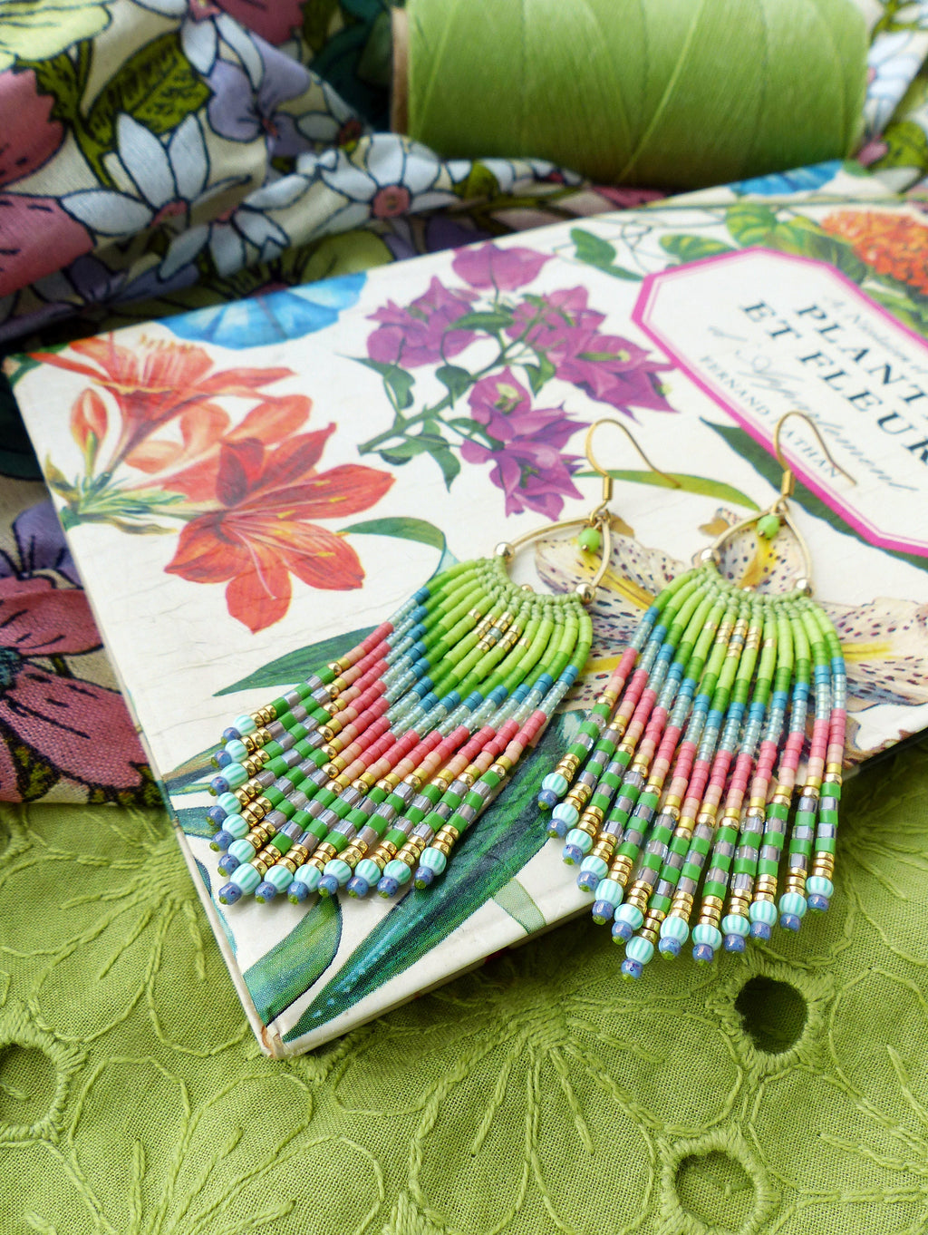 Peut inclure: Une paire de boucles d'oreilles à franges de perles colorées avec des accents dorés. Les boucles d'oreilles sont suspendues à un livre avec un motif floral et le texte "Plants et Fleurs".