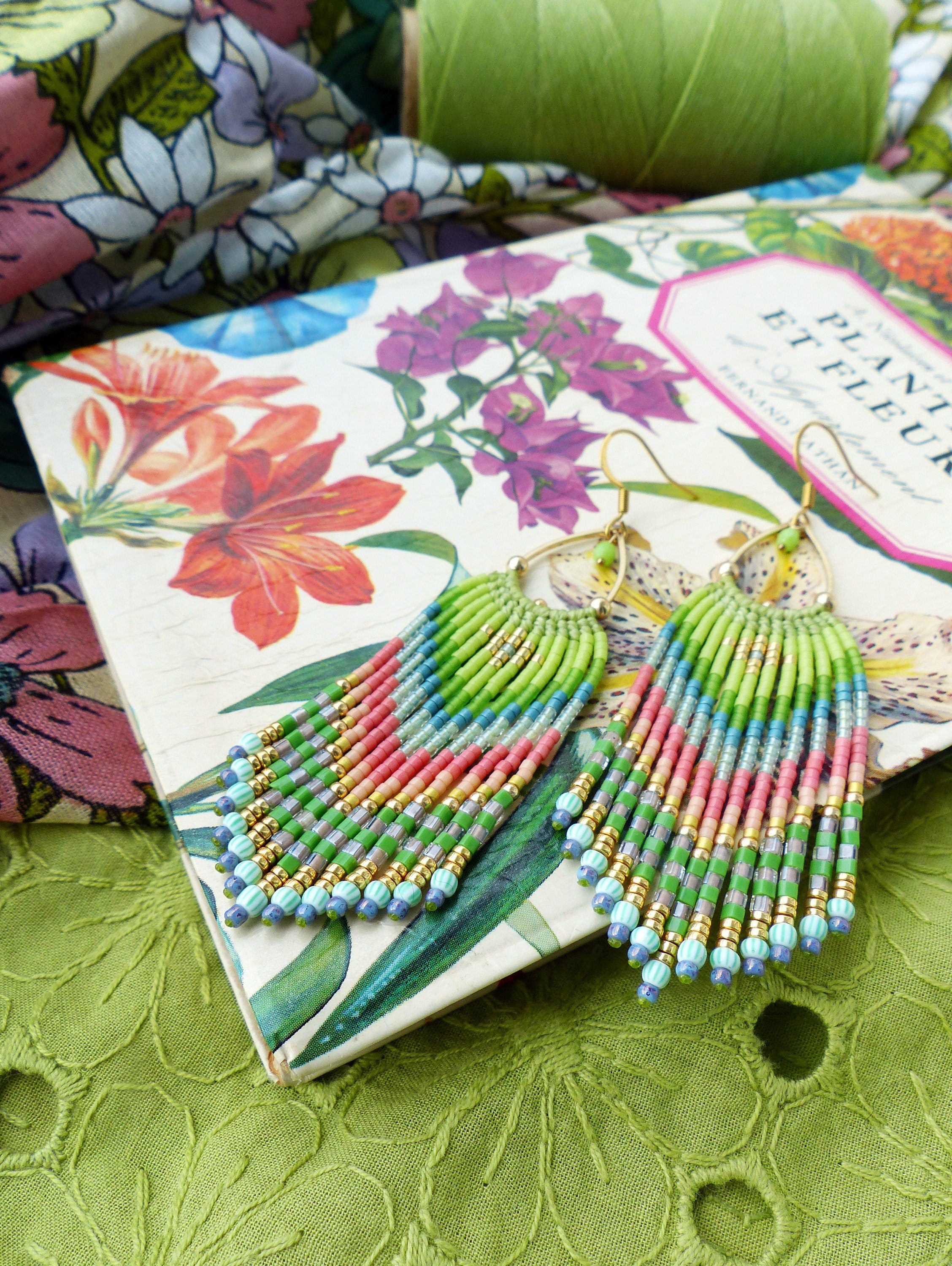 Peut inclure: Une paire de boucles d'oreilles à franges de perles colorées avec des accents dorés. Les boucles d'oreilles sont suspendues à un livre avec un motif floral et le texte "Plants et Fleurs".