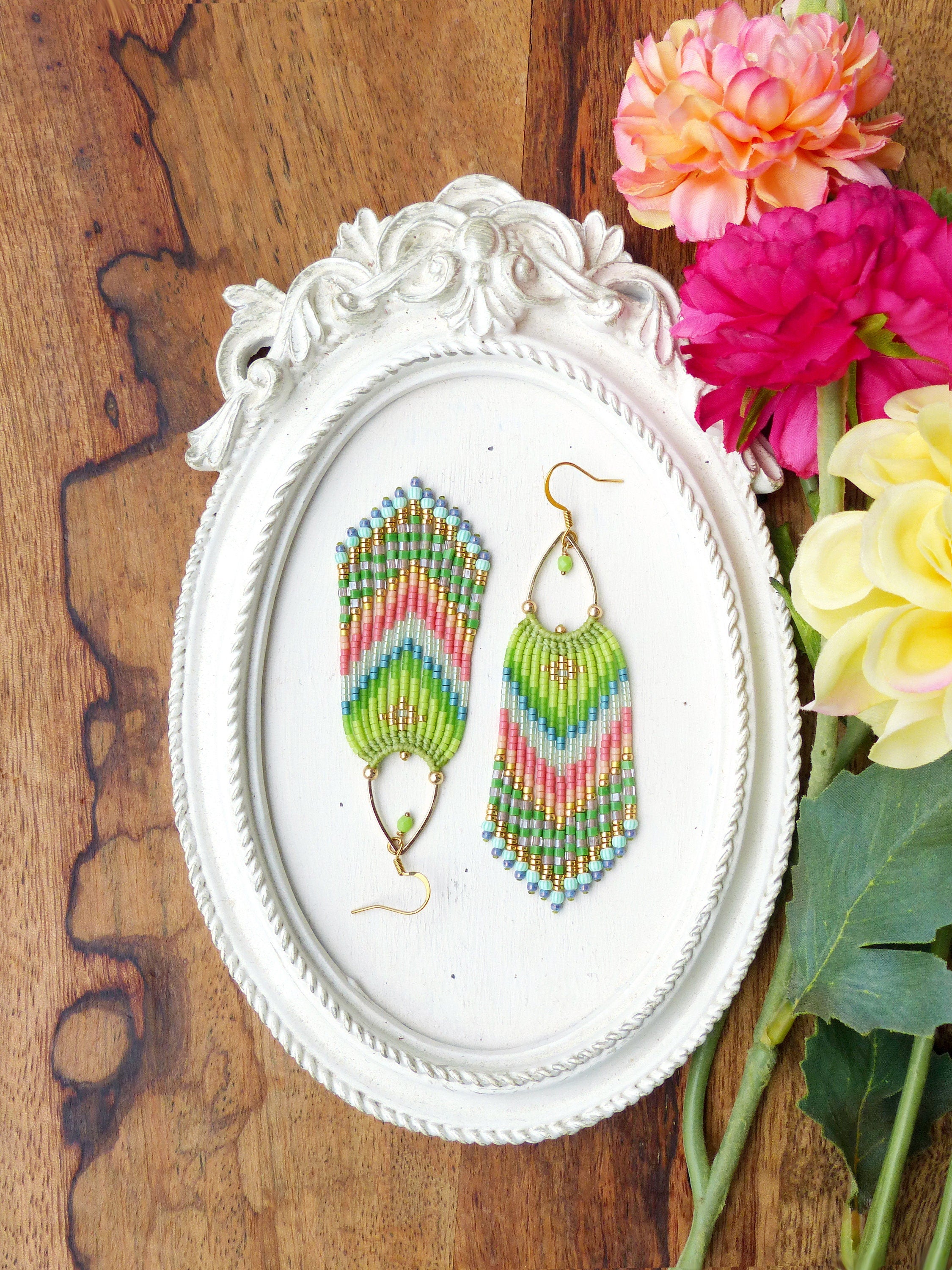 Peut inclure: Une paire de longues boucles d'oreilles en perles colorées avec un motif en chevron dans des tons de vert, rose et bleu. Les boucles d'oreilles sont suspendues à des crochets dorés et sont exposées sur un cadre ovale blanc.
