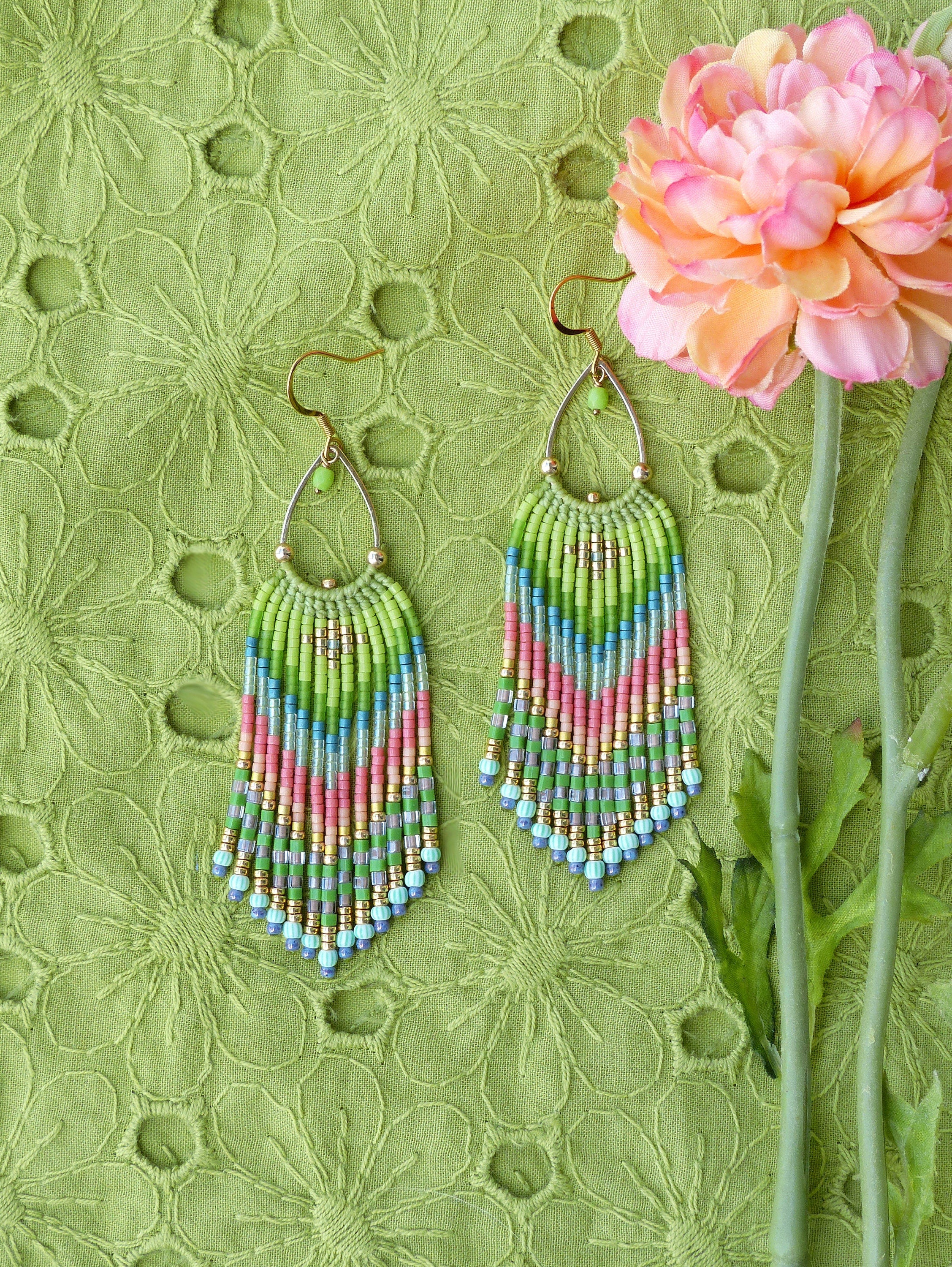 Peut inclure: Une paire de longues boucles d'oreilles pendantes en perles avec un schéma de couleurs vert, bleu et rose. Les boucles d'oreilles sont suspendues à des crochets dorés et ont une forme de goutte d'eau.