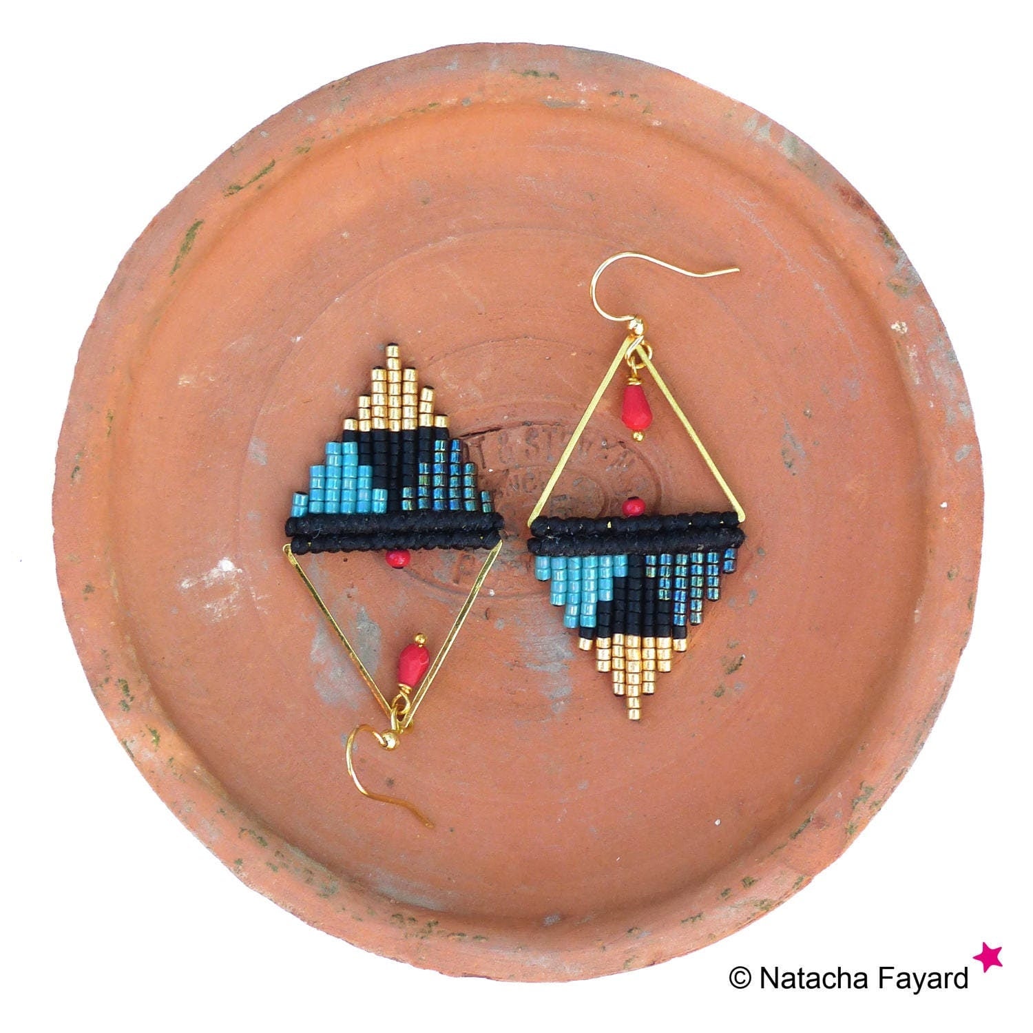 Peut inclure: Une paire de boucles d'oreilles géométriques avec un fil doré, des perles noires et bleues et des accents rouges. Les boucles d'oreilles sont présentées sur une assiette en terre cuite.