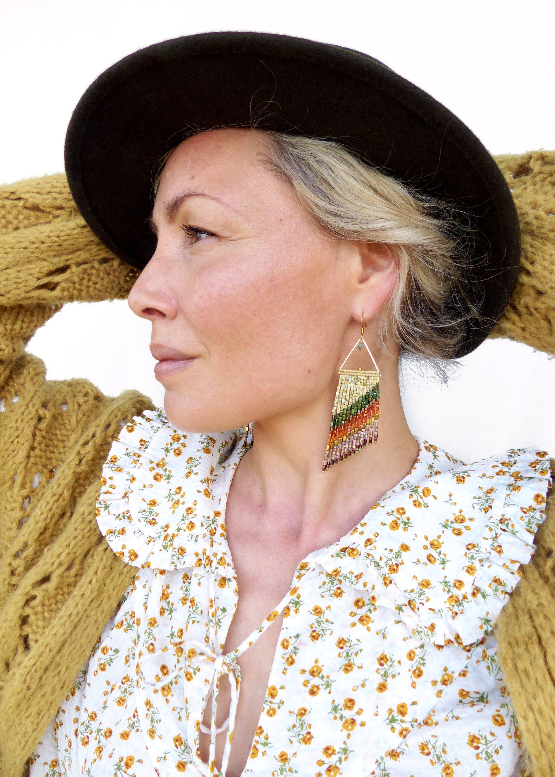 Peut inclure: Une femme portant un chapeau noir, un cardigan jaune et une blouse blanche à motif floral. Elle porte de longues boucles d'oreilles en perles de forme triangulaire et aux couleurs arc-en-ciel.