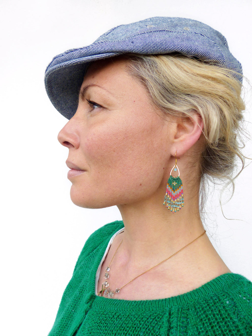 Peut inclure: Une femme portant une casquette en jean bleu et un pull vert avec un collier en chaîne dorée et des boucles d'oreilles en perles. Les boucles d'oreilles sont vertes, roses et blanches.