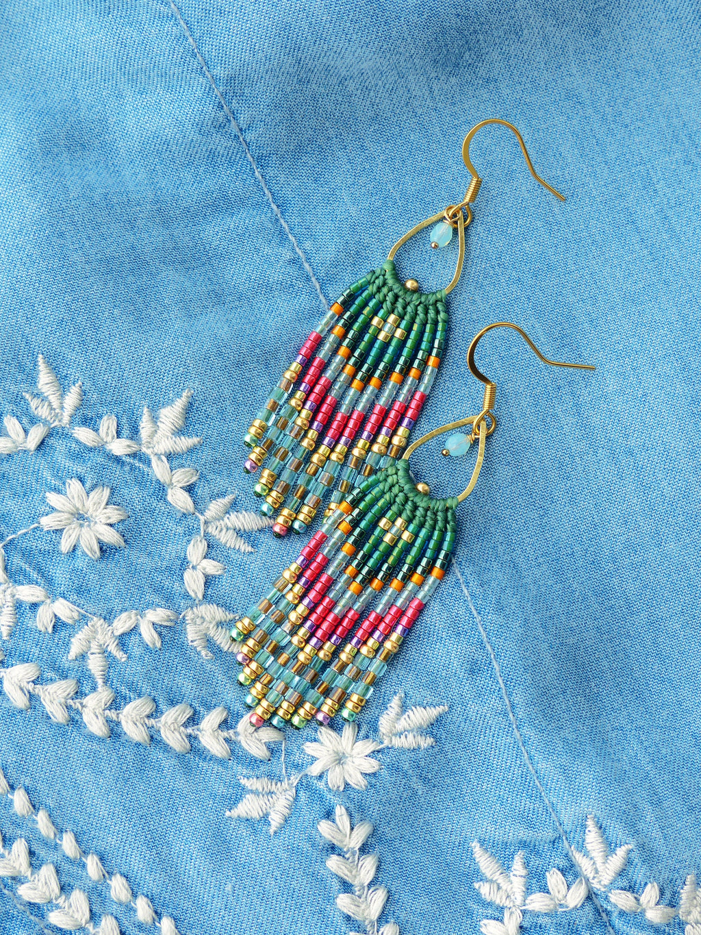 Peut inclure: Une paire de longues boucles d'oreilles en perles avec un crochet doré et un motif turquoise et rose. Les boucles d'oreilles sont suspendues sur un fond en jean bleu.
