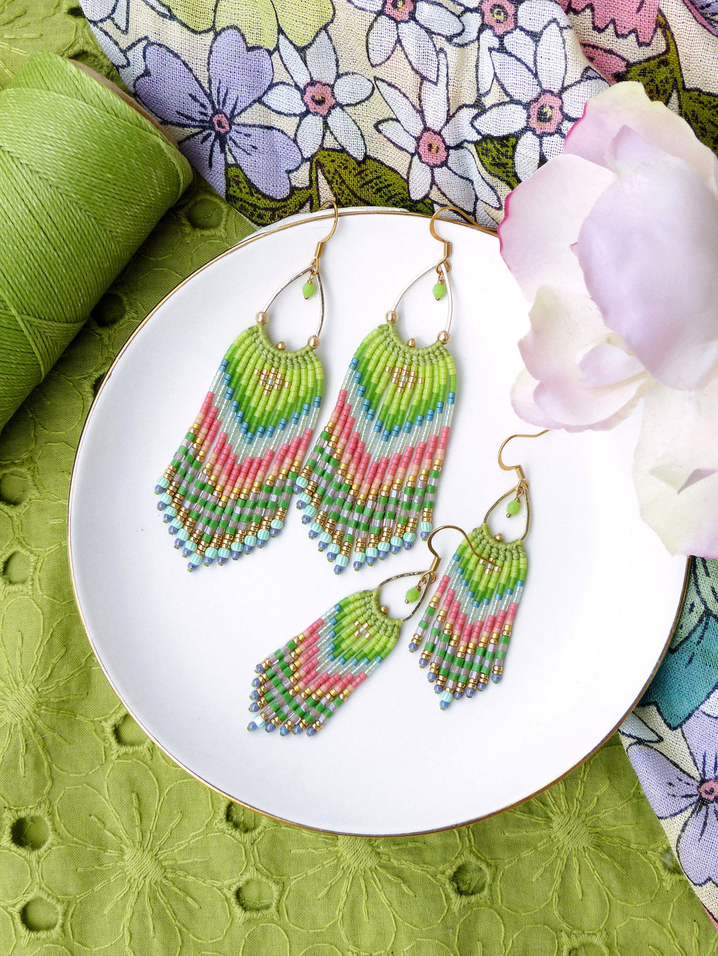 Peut inclure: Une paire de longues boucles d'oreilles en perles avec un schéma de couleurs vert, rose et bleu. Les boucles d'oreilles sont suspendues à des crochets dorés et sont sur une assiette blanche.