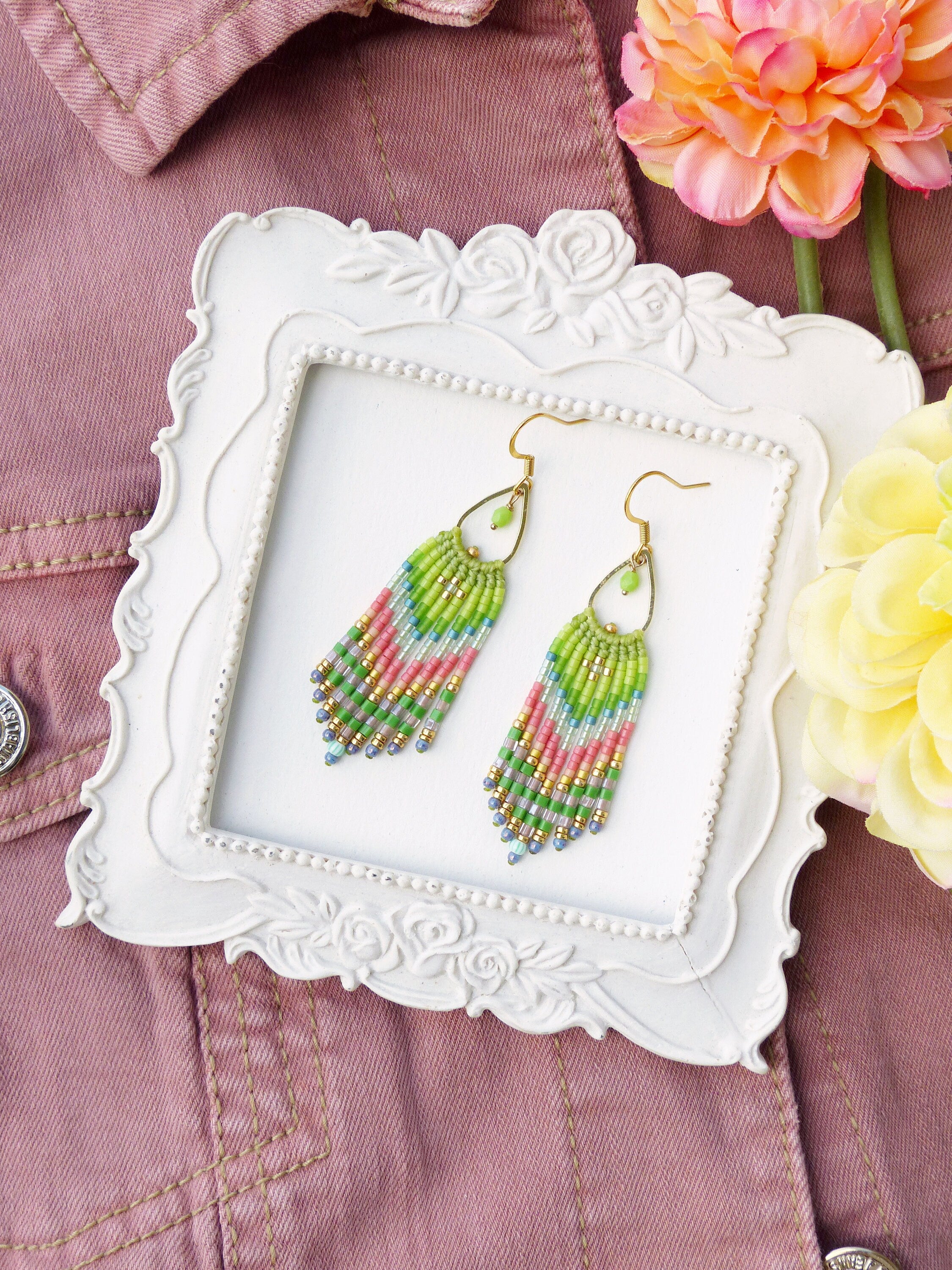 Peut inclure: Une paire de boucles d'oreilles pendantes en perles vertes, roses et bleues avec des crochets dorés. Les boucles d'oreilles sont présentées dans un cadre blanc à motif floral.
