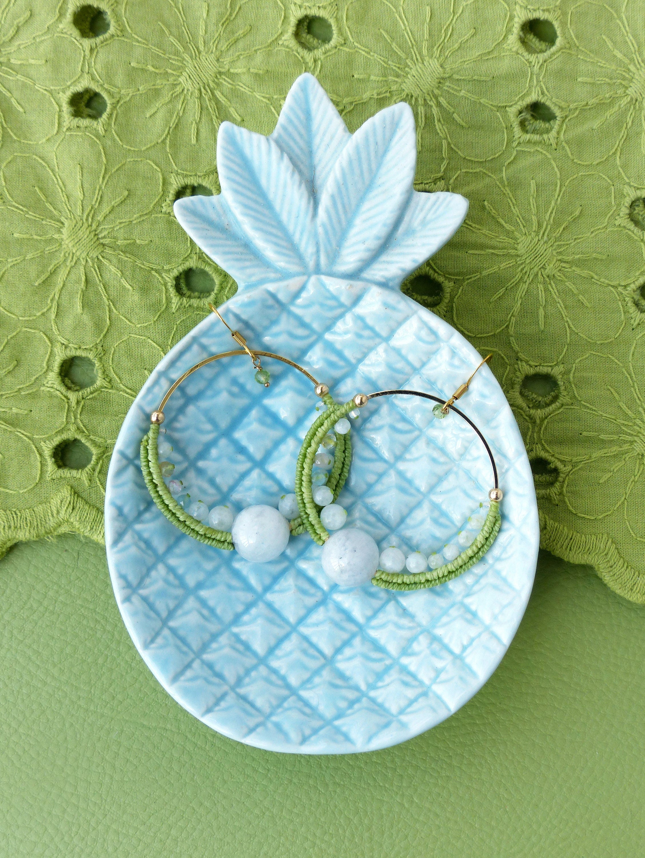 Peut inclure: Une paire de boucles d'oreilles créoles avec des perles vertes et bleues et un pendentif en pierre bleu clair. Les boucles d'oreilles sont présentées sur un plat en céramique bleu en forme d'ananas.