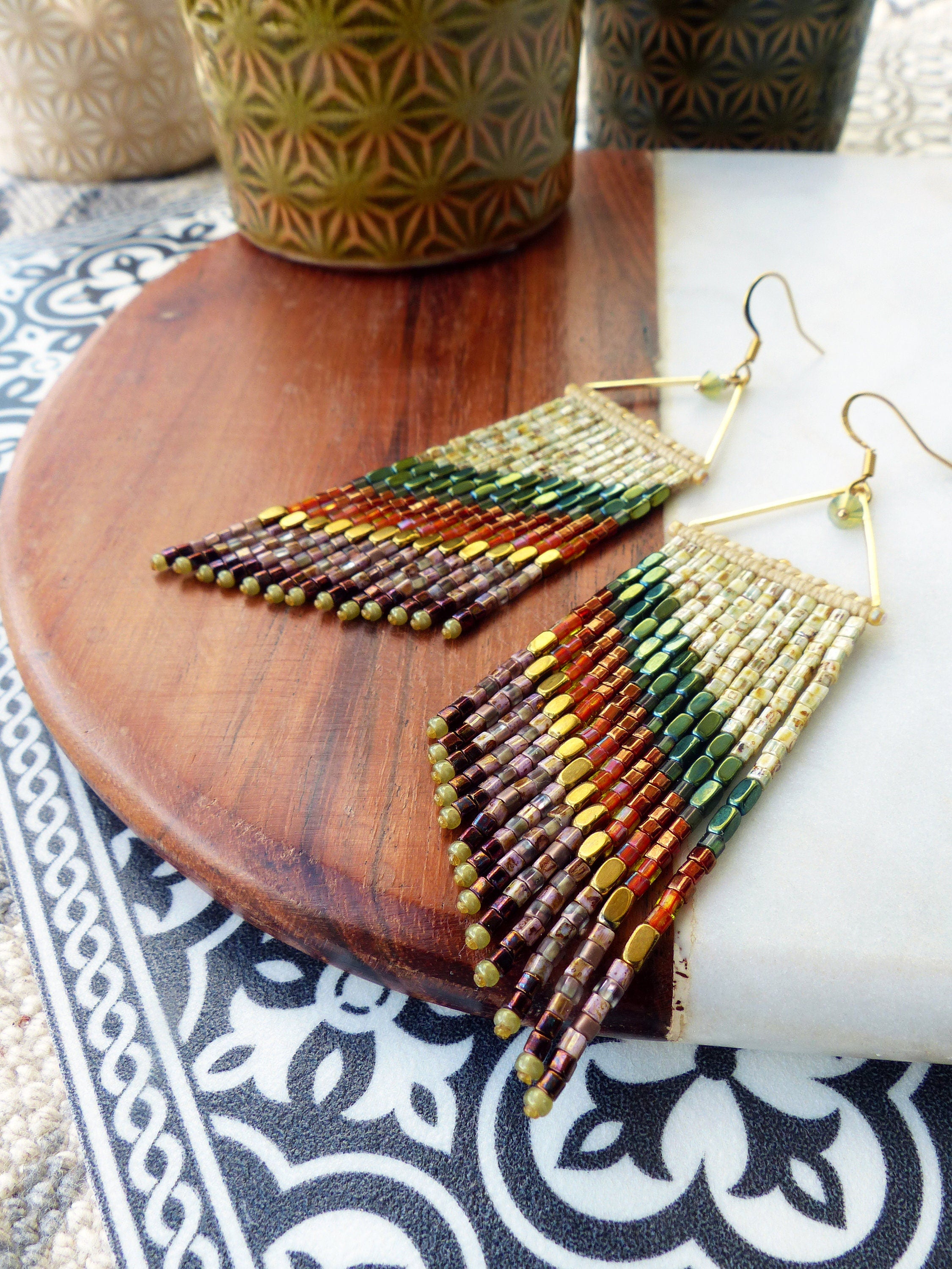 Peut inclure: Une paire de longues boucles d'oreilles en perles avec une forme triangulaire dorée en haut. Les perles sont dans des tons de brun, vert, or et blanc, créant un effet dégradé en couches.