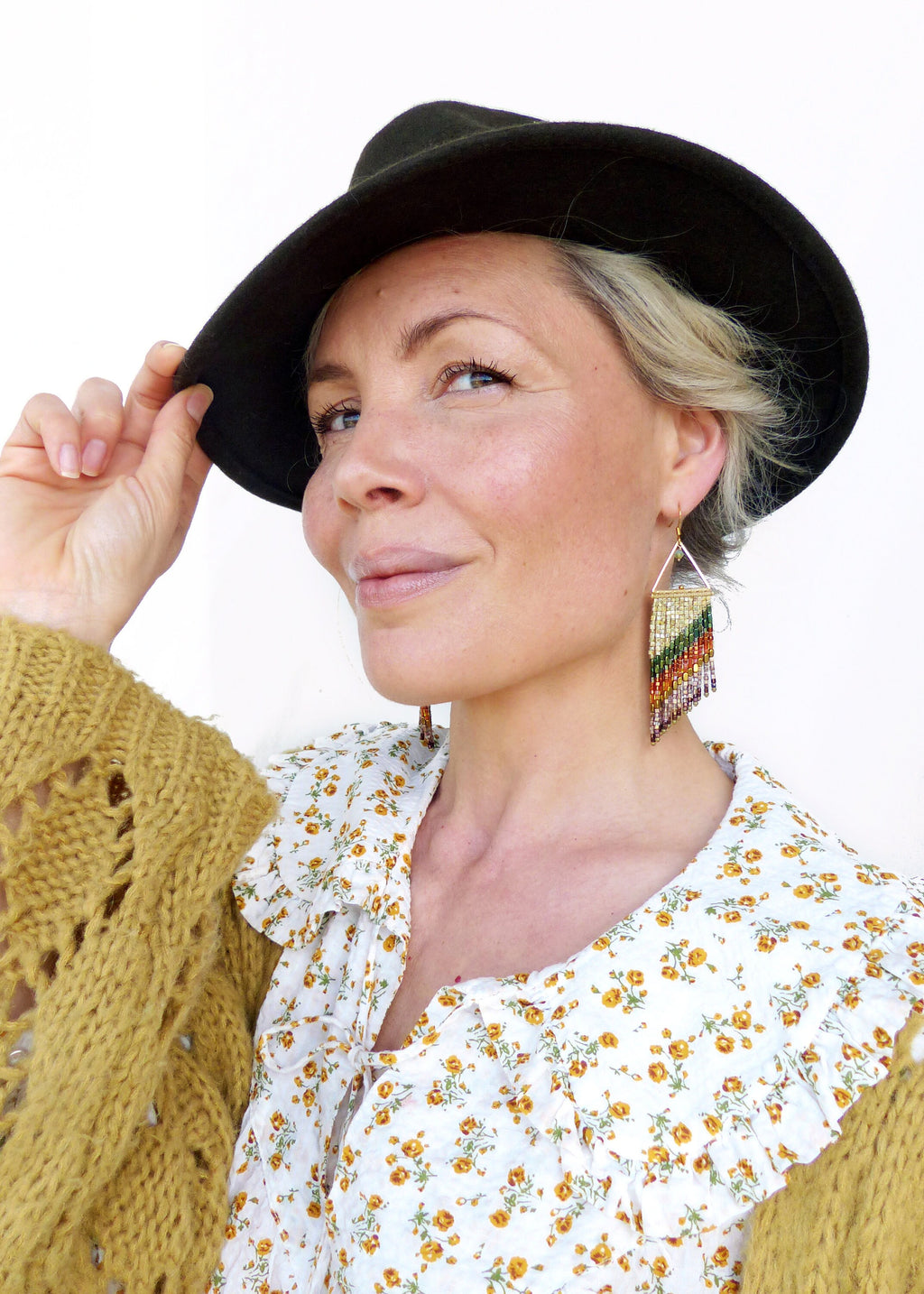 Peut inclure: Une femme portant un chapeau fedora noir et une blouse blanche à motif floral. Elle porte également de longues boucles d'oreilles en perles colorées.