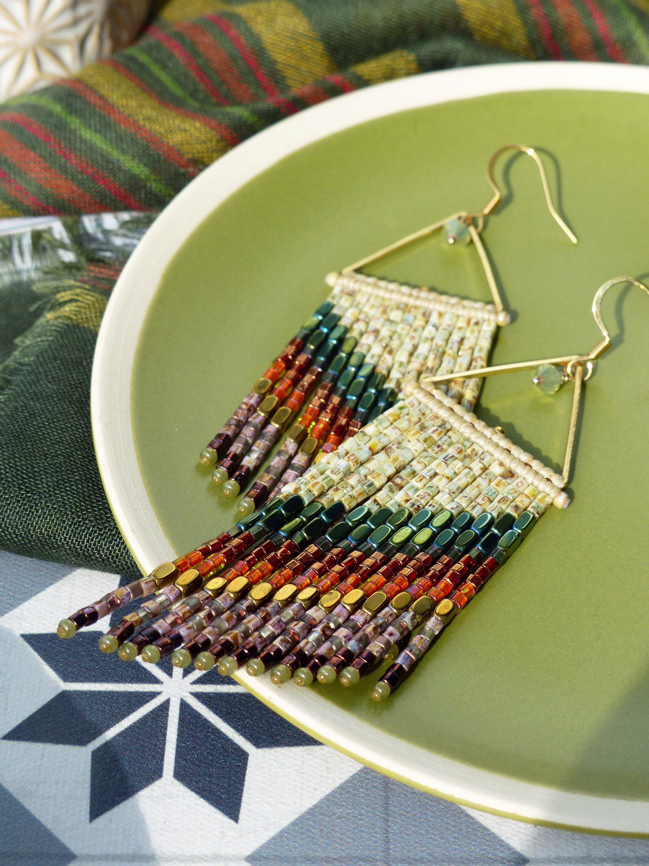 Peut inclure: Une paire de boucles d'oreilles en or avec un design triangulaire et une frange de perles colorées. Les perles sont dans les tons de vert, marron, orange et rouge.