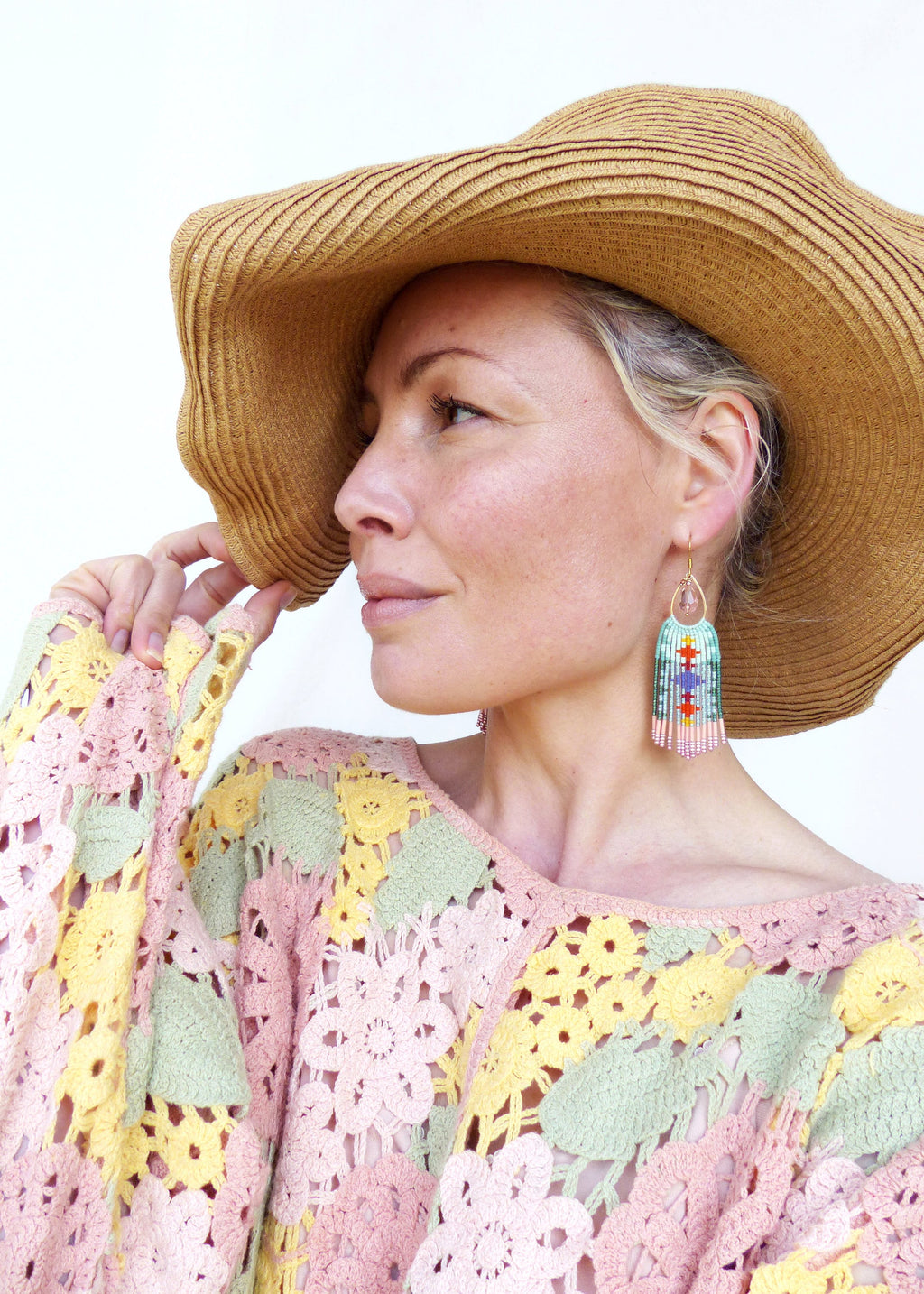 Peut inclure: Une femme porte des boucles d'oreilles dorées à franges perlées colorées. Elle porte également un chapeau de paille à larges bords et un pull en crochet pastel avec des motifs floraux roses, jaunes et verts.