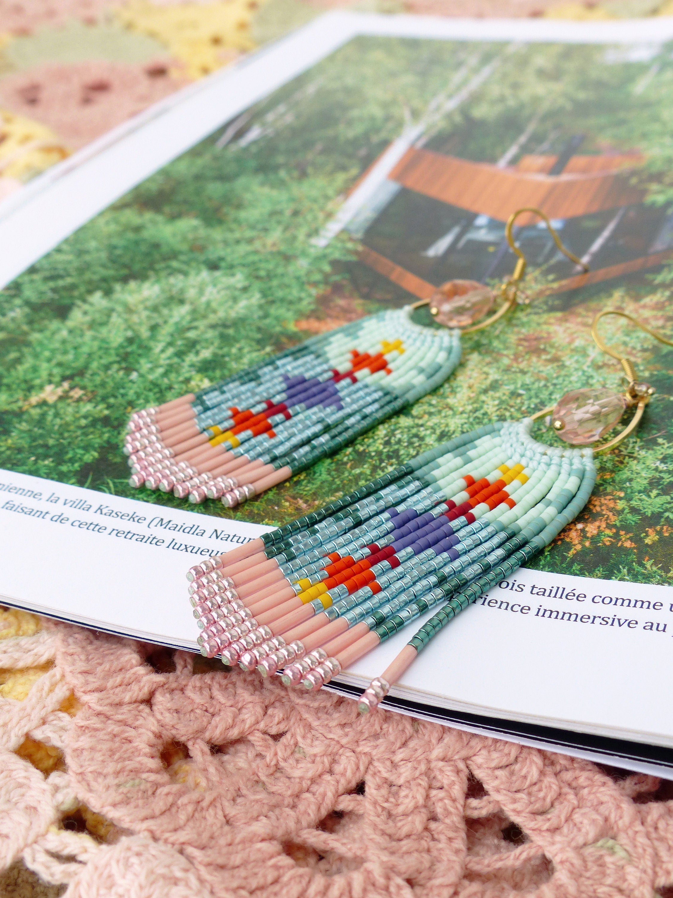 Peut inclure: Boucles d'oreilles en perles avec un motif de franges. Les boucles d'oreilles présentent un schéma de couleurs vert clair et rose avec des accents de violet, orange et jaune. Elles sont posées sur un magazine avec du texte et une couverture crochetée.