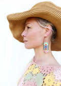 Peut inclure: Une femme porte un chapeau de paille à larges bords et des boucles d'oreilles en perles. Les boucles d'oreilles ont un crochet doré et un motif rectangulaire avec des perles bleues, vertes, rouges et roses. La femme porte un haut en crochet rose, jaune et vert.