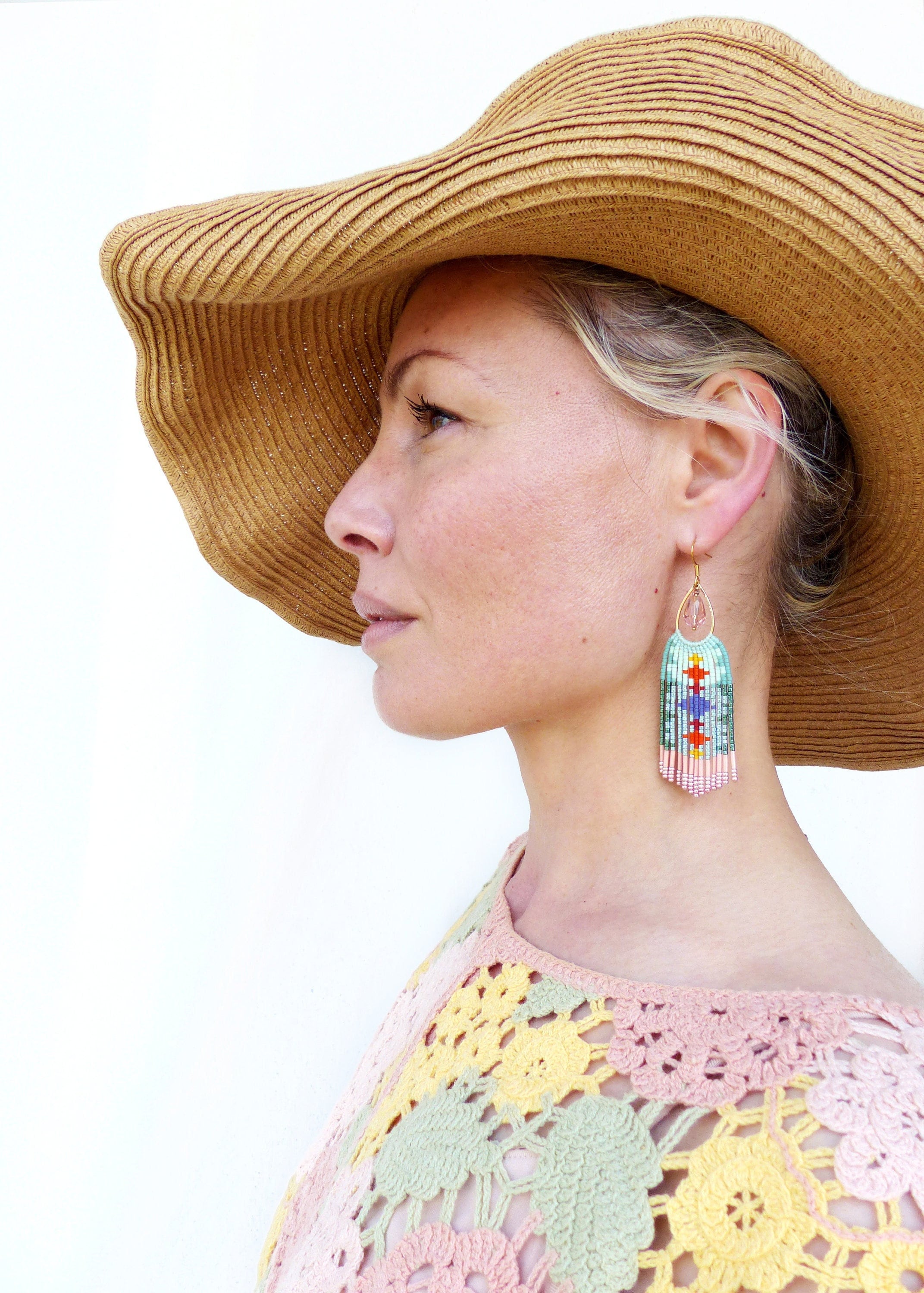 Peut inclure: Une femme porte un chapeau de paille à larges bords et des boucles d'oreilles en perles. Les boucles d'oreilles ont un crochet doré et un motif rectangulaire avec des perles bleues, vertes, rouges et roses. La femme porte un haut en crochet rose, jaune et vert.