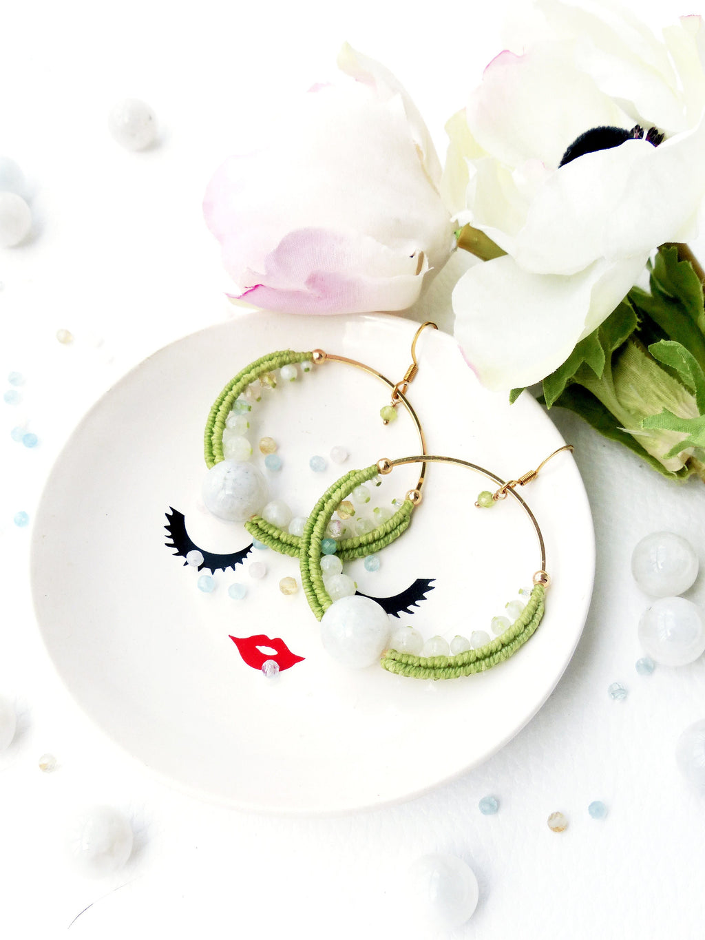 Peut inclure: Une paire de boucles d'oreilles créoles vertes et blanches avec un accent de pierre blanche. Les boucles d'oreilles sont sur une assiette blanche avec un motif de visage noir et rouge.