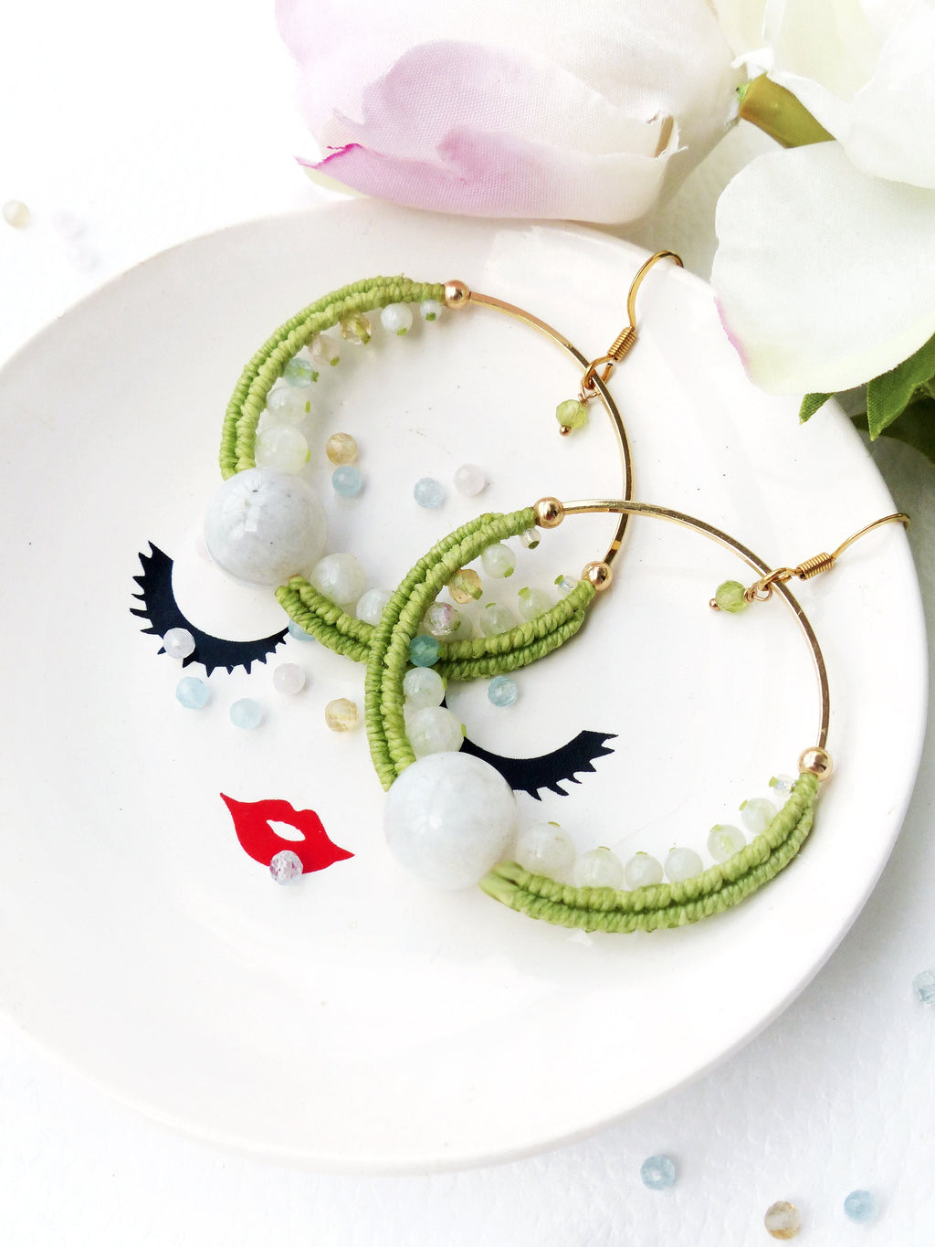 Peut inclure: Une paire de boucles d'oreilles créoles vertes et dorées avec des accents de pierres blanches. Les boucles d'oreilles sont sur une assiette blanche avec un motif de visage noir et blanc.
