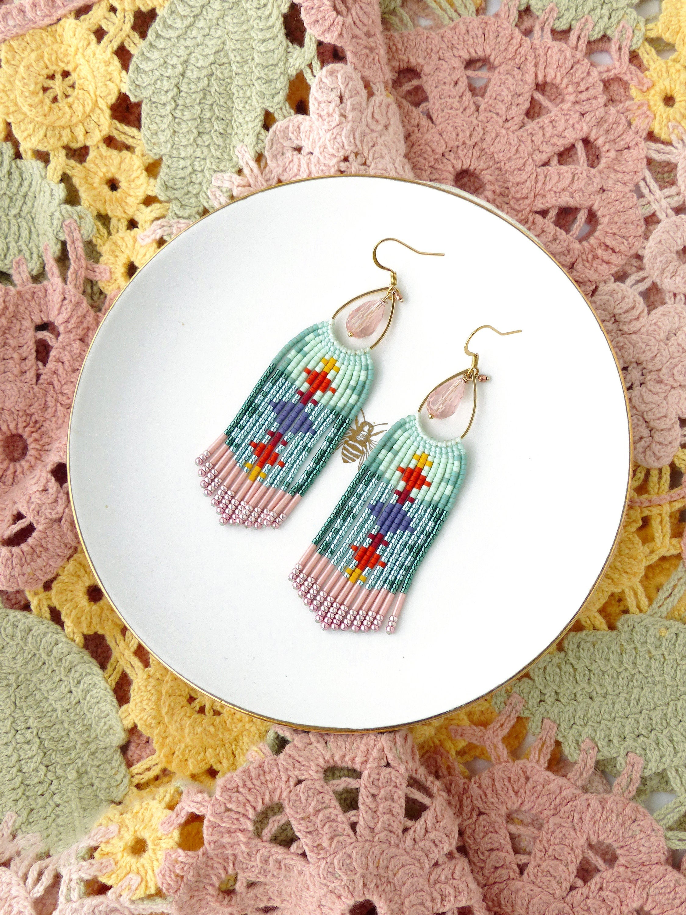 Peut inclure: Paire de boucles d'oreilles en perles avec un motif turquoise, rose, rouge, jaune et violet. Les boucles d'oreilles ont des crochets d'oreilles dorés et une perle rose en forme de goutte. Elles sont présentées sur une assiette blanche.