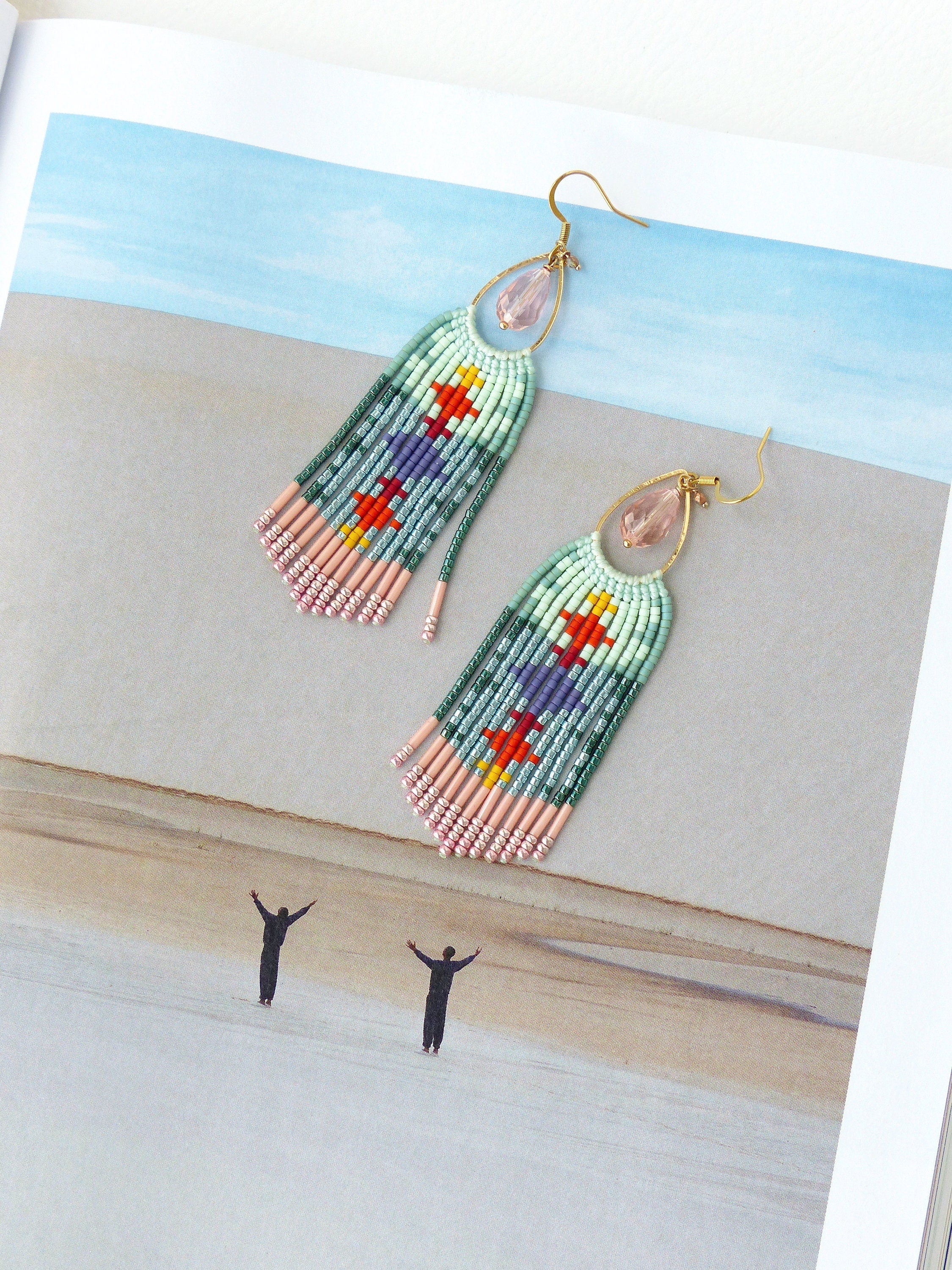 Peut inclure: Une paire de boucles d'oreilles en perles avec un motif géométrique coloré. Les boucles d'oreilles comportent un crochet doré et une perle rose en forme de goutte. Le corps principal des boucles d'oreilles est rectangulaire avec une frange de perles roses en bas.