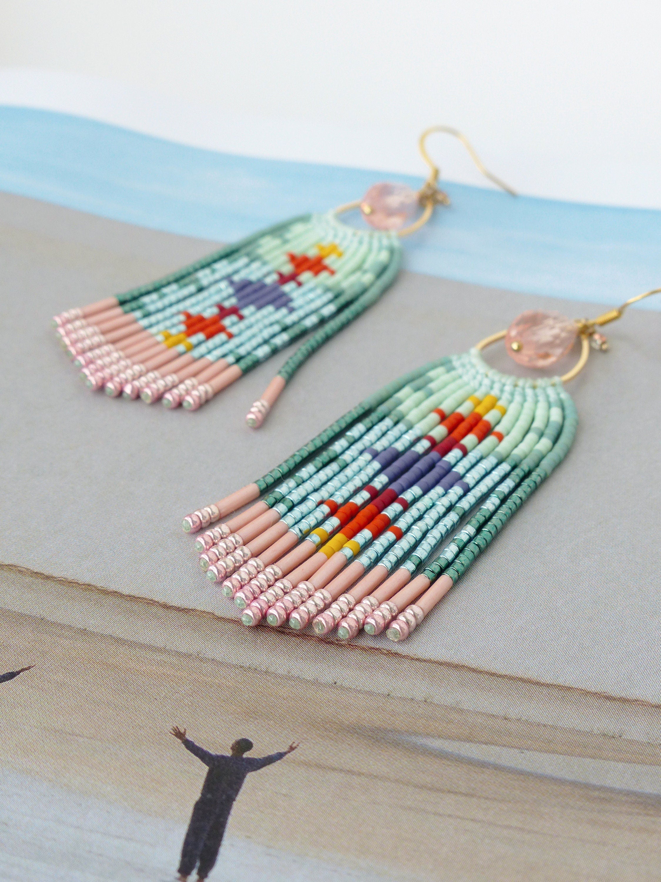 Peut inclure: Boucles d'oreilles faites à la main avec un motif géométrique coloré. Les boucles d'oreilles présentent une frange de perles vert clair, roses et bleues, avec des accents de rouge, de jaune et de violet. Chaque boucle d'oreille a un crochet doré et une pierre rose.
